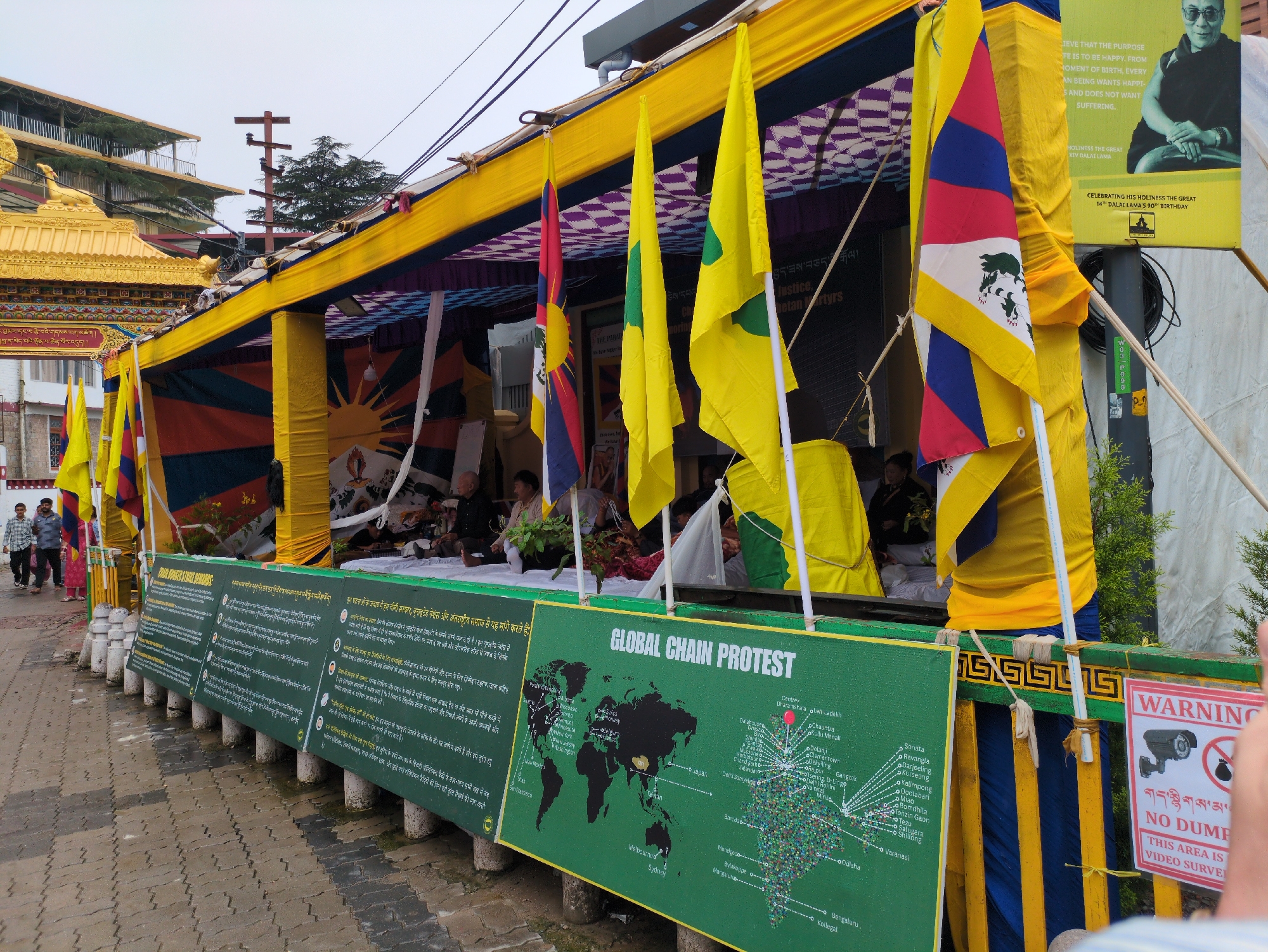 This image depicts a street scene in Dharamshala, India, likely during a protest event. The foreground is dominated by a long, green banner with the text "GLOBAL CHAIN PROTEST" and a world map marked with various locations. The banner is supported by white pillars and a yellow and blue striped fabric structure.
Behind the banner, a temporary yellow canopy stretches across the scene, adorned with purple and white checkered fabric on the underside. Several flags are visible, including a Tibetan flag with its distinctive imagery of a sun, mountains, and snow lions, as well as plain yellow flags with green symbols. A large poster featuring a portrait of the Dalai Lama and text in English and Tibetan is prominently displayed on the right side of the canopy.
Several people are present. In the middle ground, individuals are seated or standing behind the banner, some appearing to be participants or organizers of the event. In the background, to the left, two individuals are walking away from the camera on a paved walkway. The architecture in the background suggests a hillside setting, consistent with Dharamshala's topography. The sky is overcast, indicating potentially cool or rainy weather. A "WARNING NO DUMPING THIS AREA IS UNDER VIDEO SURVEILLANCE" sign is visible on the right edge of the frame.