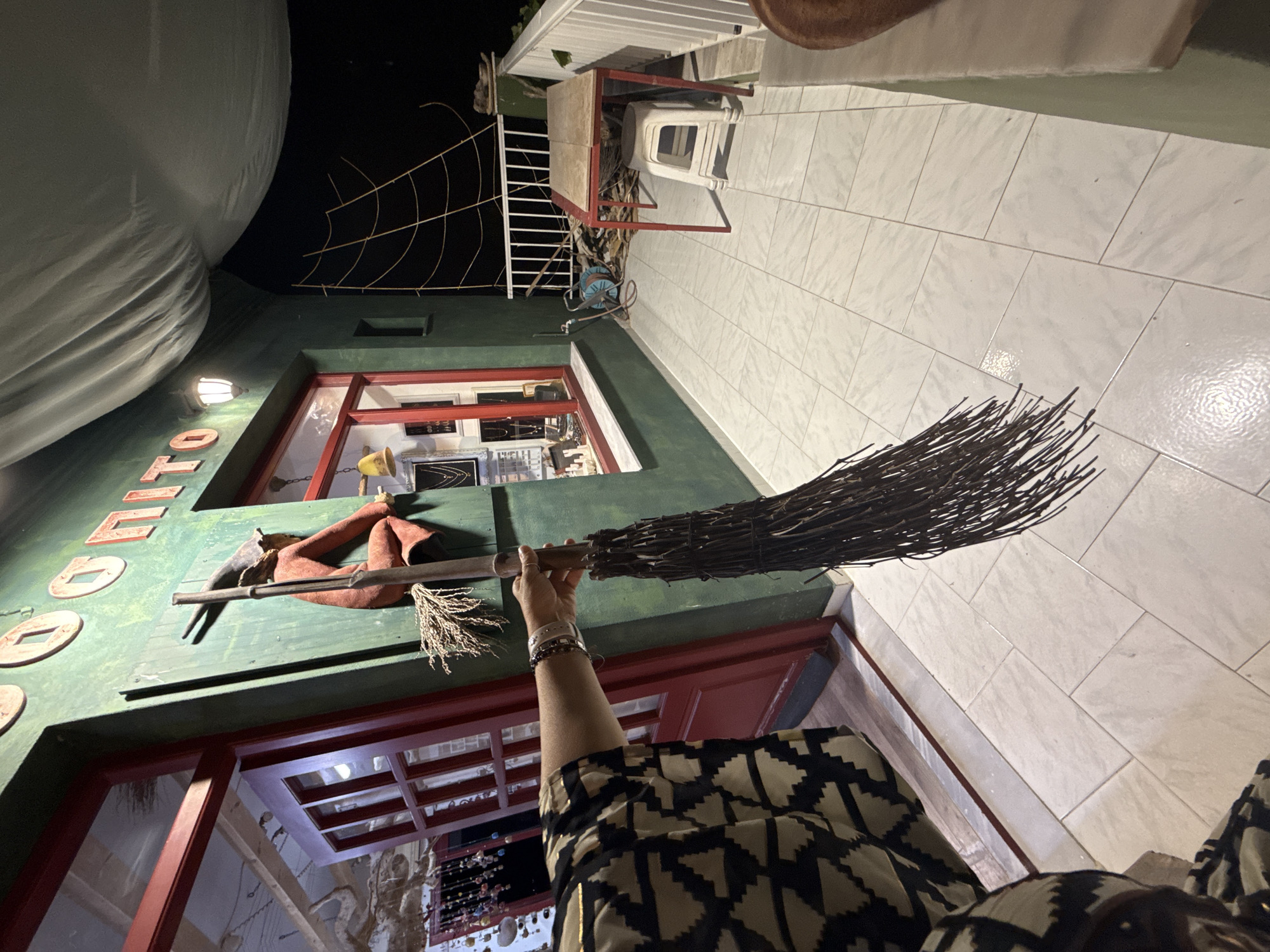 A person's arm, adorned with bracelets, holds a rustic broom made of dark twigs. The broom is positioned in the foreground, extending from the right edge of the frame towards the left. The person is wearing a black and tan patterned garment. In the background, a green building with red trim features a window displaying jewelry. Above the window, large, round, light-colored letters form a word on the building's facade. A decorative element of a witch figure on a broomstick is mounted on the green wall. To the right of the building, a white railing encloses an outdoor area with a tiled floor. A red-legged table with a light-colored wooden top sits on the tiled floor, accompanied by a white plastic stool. A coiled blue hose and other debris are visible near the table. The scene is dimly lit, suggesting it is nighttime. This appears to be an outdoor space in front of a shop, possibly in the village of Pelekas, Greece, given the architectural style and the hint of a spiderweb decoration on the wall.