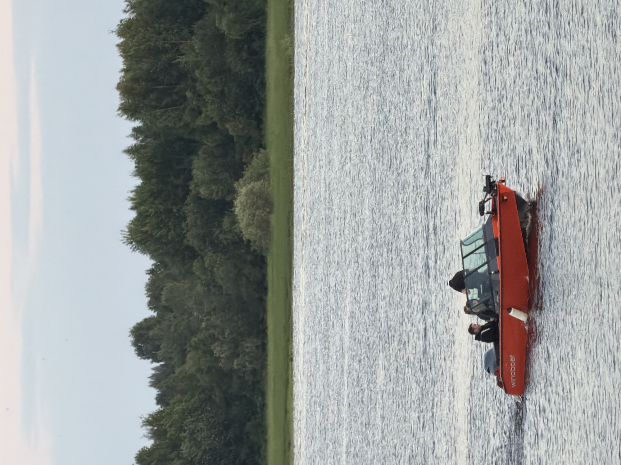 An orange motorboat with three people aboard is traveling on a wide body of water. The boat has a black windshield and trim. One person appears to be at the helm, while two others are seated. The boat's hull is emblazoned with the word "windboat" in white lettering. The water is choppy, with ripples and small waves. In the background, a dense line of green trees forms the shoreline. The sky above is a pale blue with thin, wispy clouds. The scene appears to be a serene moment on a lake or river, possibly during daylight hours. The location is not identifiable within the city of Solikamsk, Russia, based on the provided image.