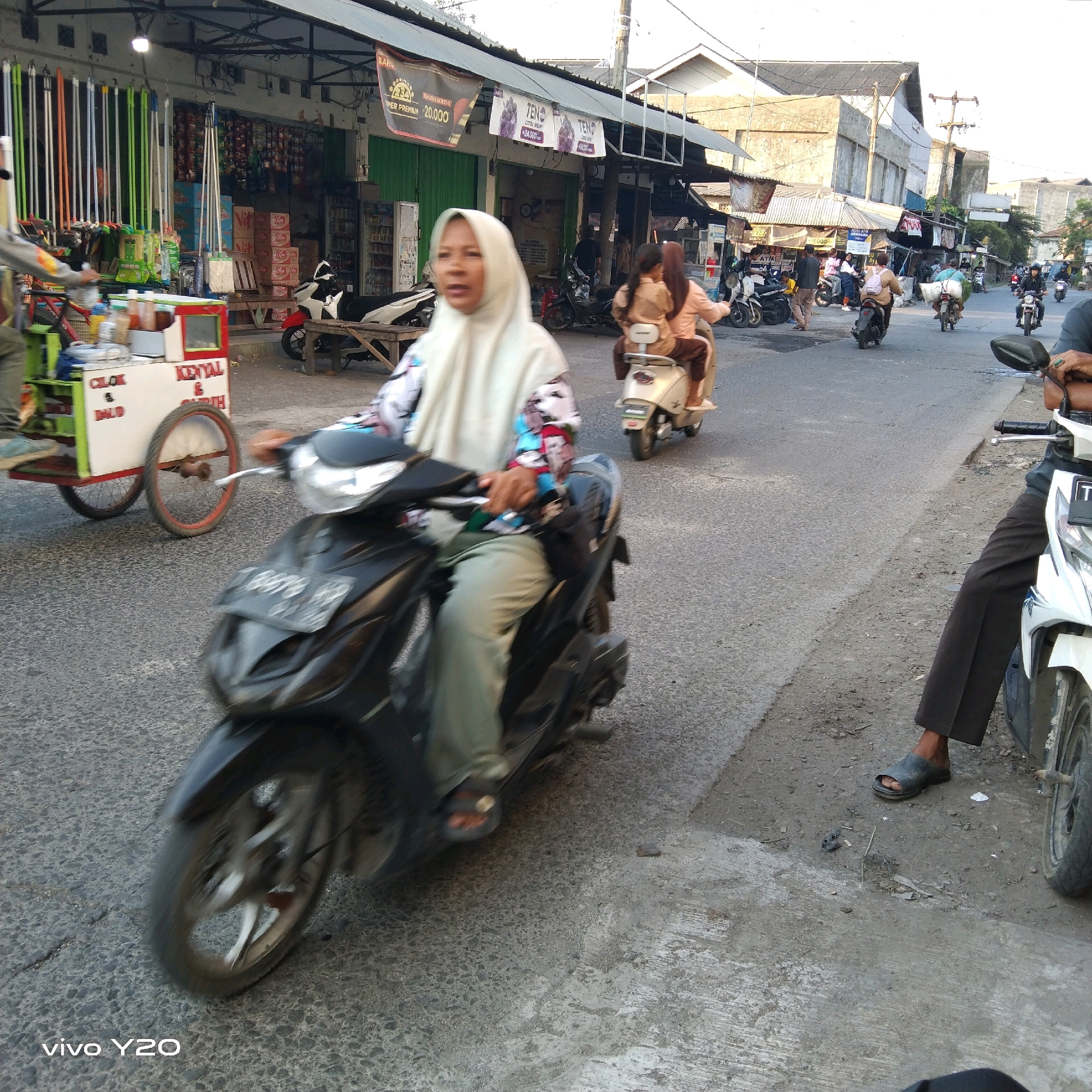 This image depicts a street scene in Karawang, Indonesia, likely during daylight hours given the lighting. In the foreground, a person is riding a black motorcycle, wearing a white hijab and a colorful patterned top, with light green pants and sandals. The motorcycle's front tire and part of its license plate are visible. To the left of the motorcycle, a food cart with red and green accents is being pulled. The cart has text that reads "CILOK & DAUD KENYAL & GURIH". In the background, several other motorcycles and people are present on the street, some riding and others standing. Along the side of the street are storefronts with awnings, displaying goods such as brooms and various packaged items. One storefront has banners with prices indicated. The environment appears to be a busy urban street with commercial activity.