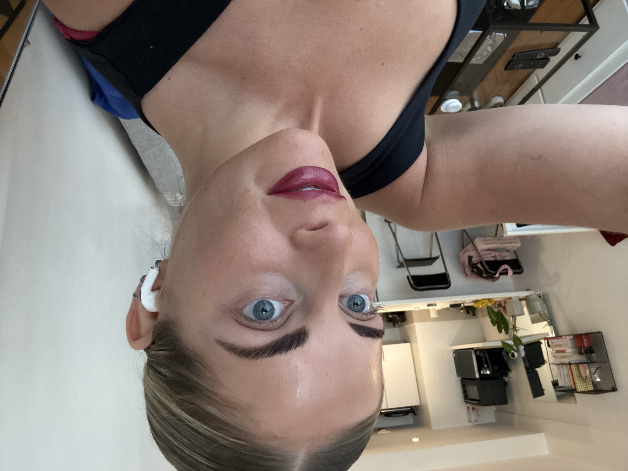 A woman is looking at the camera. She is wearing a black tank top and has blue eyes and dark pink lipstick. She is wearing a white Bluetooth earpiece. The background shows a kitchen setting with white cabinets and appliances, including a microwave and a wire rack filled with items. There is also a plant and a hanging wire rack above the counter. In the upper background, there is a ladder-style rack and a bag on a shelf. The location is likely an apartment or home kitchen, but there's no information to determine the city of Paris.