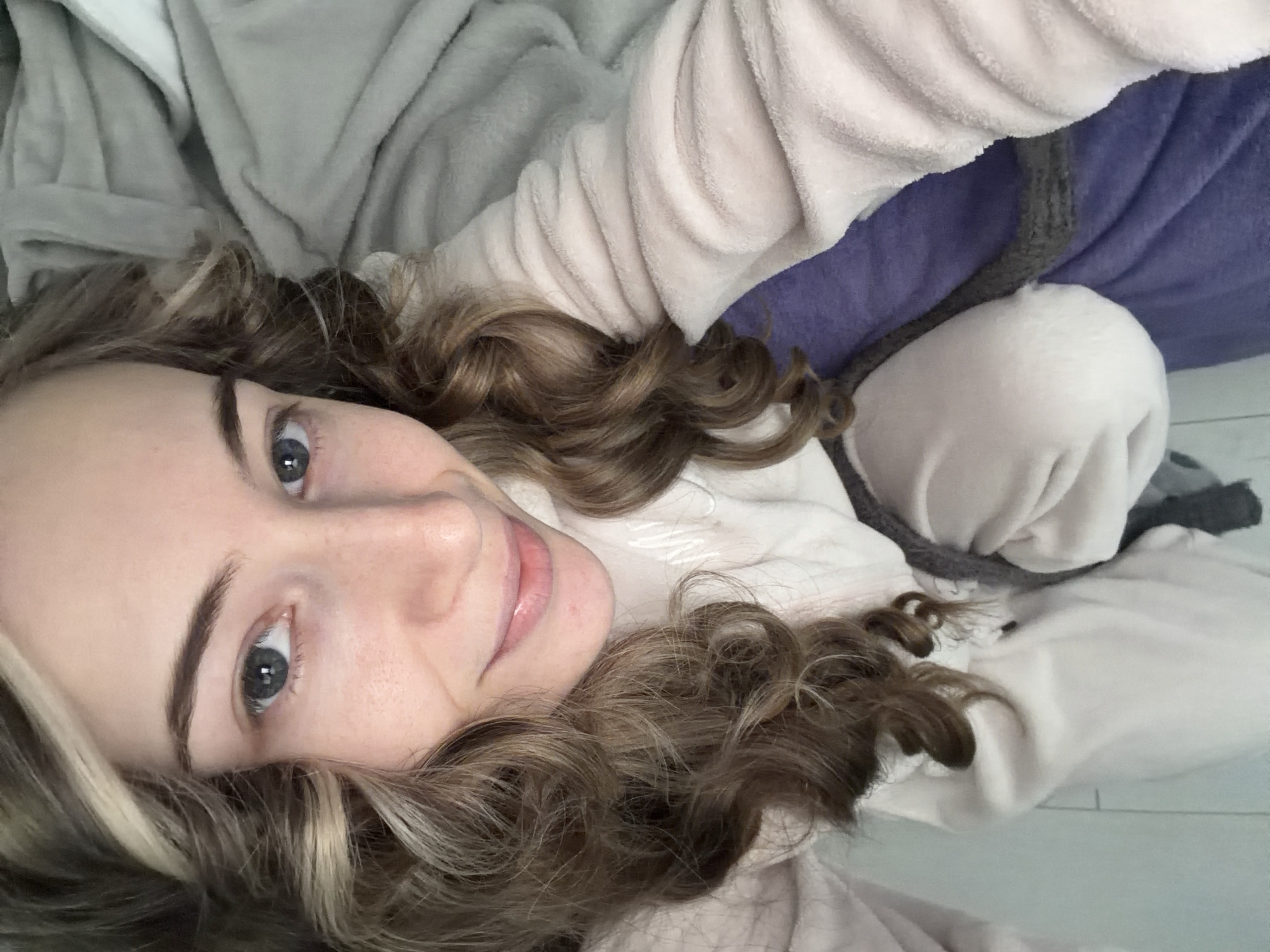 A selfie shot from a slightly high angle shows a fair-skinned woman with wavy, light brown hair. Her head rests on a light-colored, possibly white or beige, soft fabric, likely part of clothing or bedding. A small portion of gray fabric can be seen on the left side. To the right, there is a purple cushion or piece of furniture with a gray rope or cord-like object extending across it. The background suggests an indoor setting, possibly a bedroom. There is no information to determine if the scene is located in Plovdiv, Bulgaria.