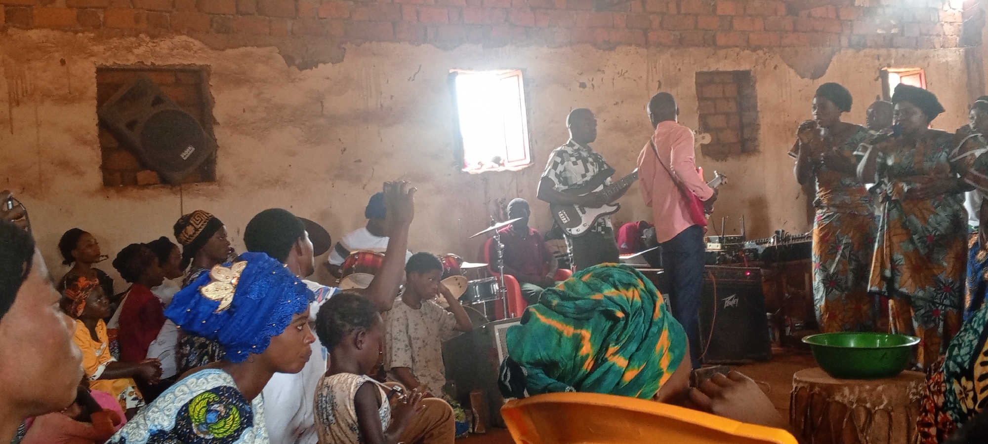 The image depicts a gathering of people, likely in Ngalula, Democratic Republic of the Congo, inside a building with a red brick wall structure and visible light coming through the windows. A band is set up with drums, bass guitar, electric guitar, and a sound system with a speaker placed in a niche in the wall. There are several women dressed in traditional African patterned clothing singing into microphones. In front of the band, a group of people is seated and watching the performance. In the foreground, the back of chairs and seated individuals are visible, providing a sense of being part of the audience. The general atmosphere is one of cultural celebration or worship. A green bowl is placed on top of a drum.
