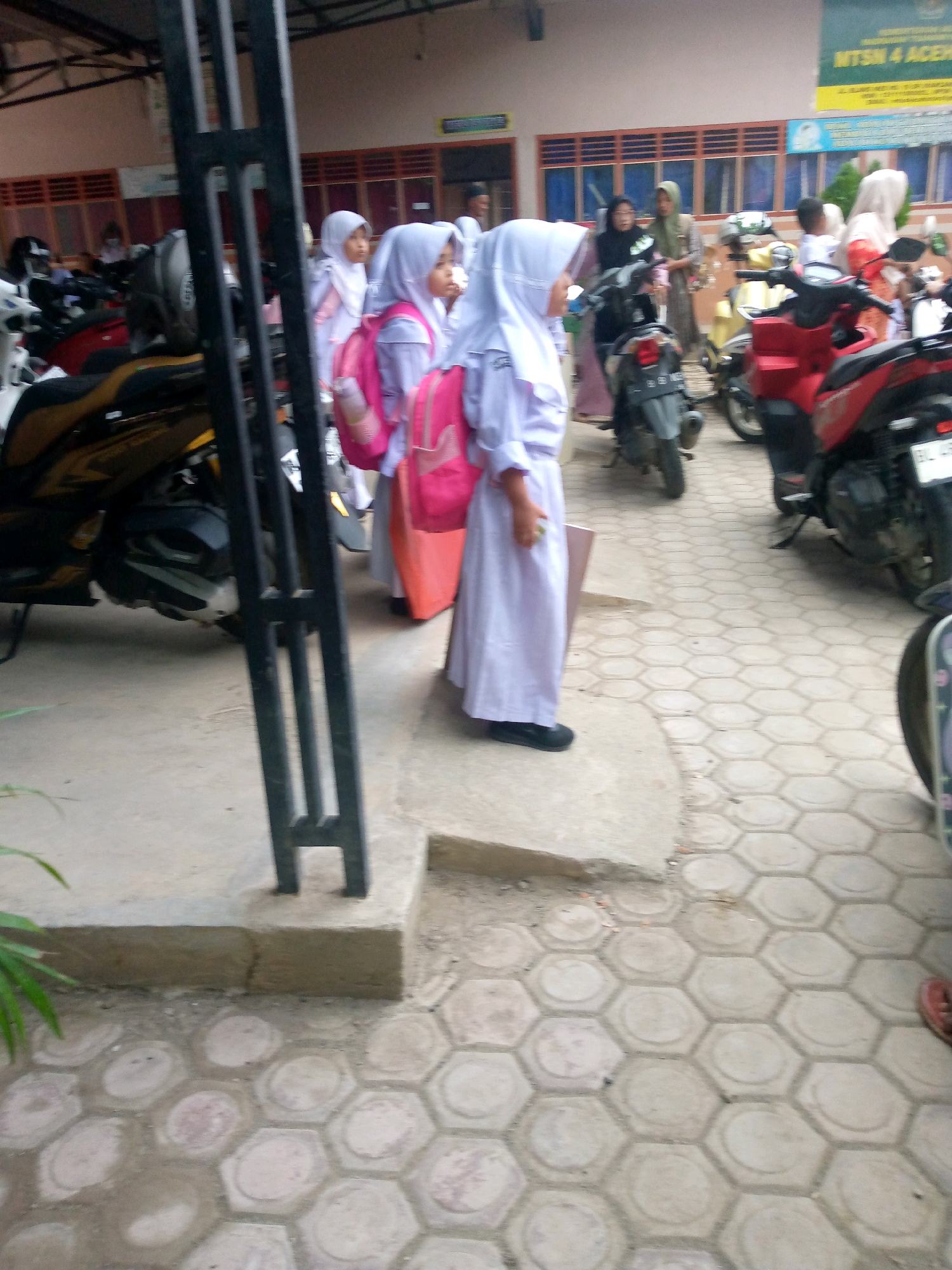 The image depicts a street scene in front of a building, likely a school, in the city of Mesjid, Indonesia. The foreground shows a concrete sidewalk with hexagonal patterned paving stones and a small plant. To the left, a black metal fence-like structure is visible. A parked motorcycle with a yellow and black body is partially visible.
In the middle ground, several students, appearing to be young girls, are walking. They are wearing light-colored hijabs and uniforms, with some carrying pink backpacks. They are seen walking past parked motorcycles. To the right, a red motorcycle is parked. The building behind the students has a pinkish-brown facade with windows and a sign at the top that reads "MTSN 4 ACEH".
In the background, more people are visible, some possibly parents or teachers, standing near other parked motorcycles. The overall environment suggests a busy area, possibly during school dismissal or arrival.