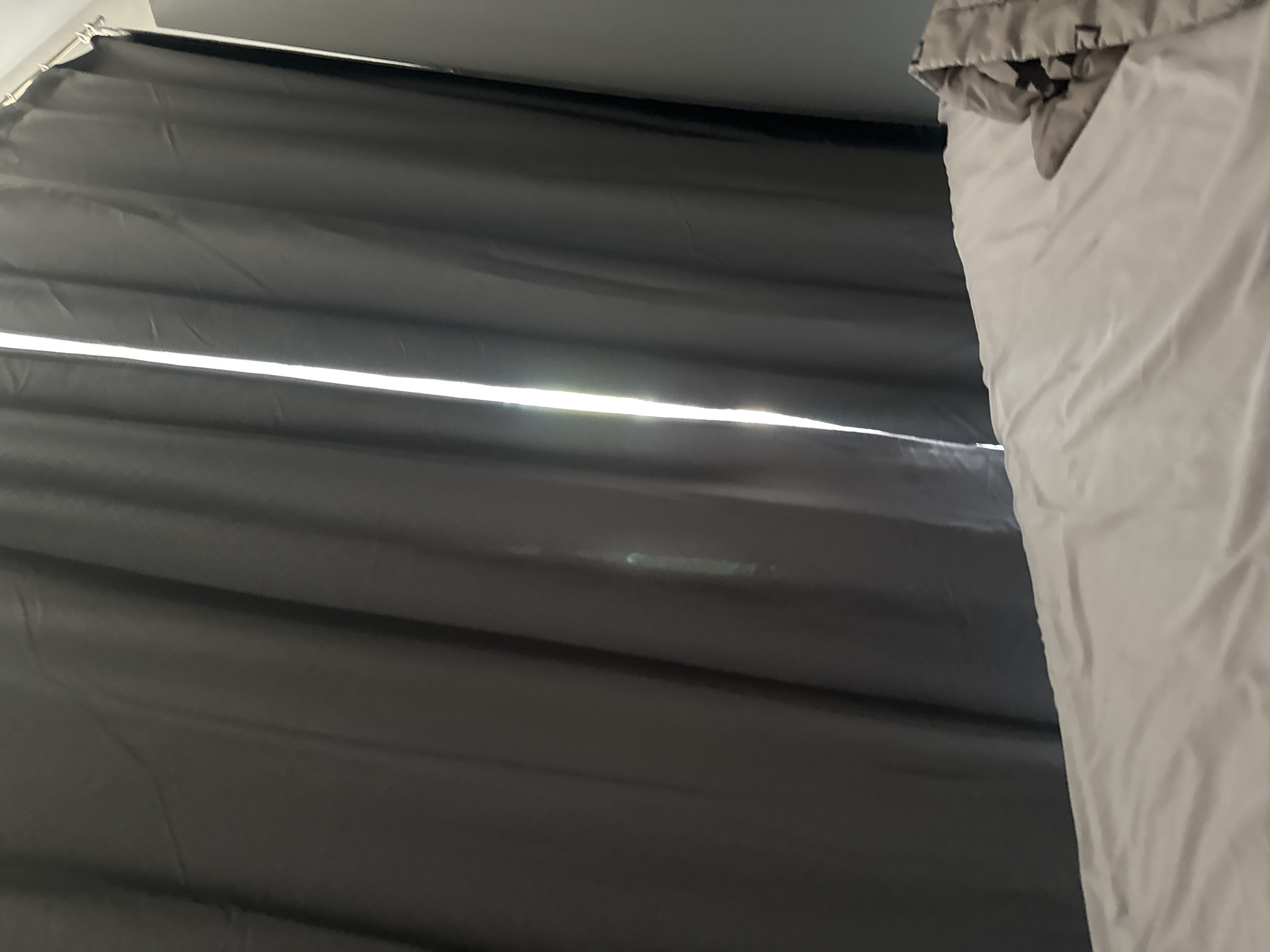 A low-angle shot shows a partial view of a room. To the left, heavy, dark gray curtains hang, allowing a horizontal sliver of bright light to penetrate through a gap. To the right, a light gray, possibly white, duvet or comforter is partially visible, with a section folded over at the top. The scene is simply a view of these items, without any apparent people, actions, or interactions taking place. The location Бобровский, Russia is not discernable from the details in this image.