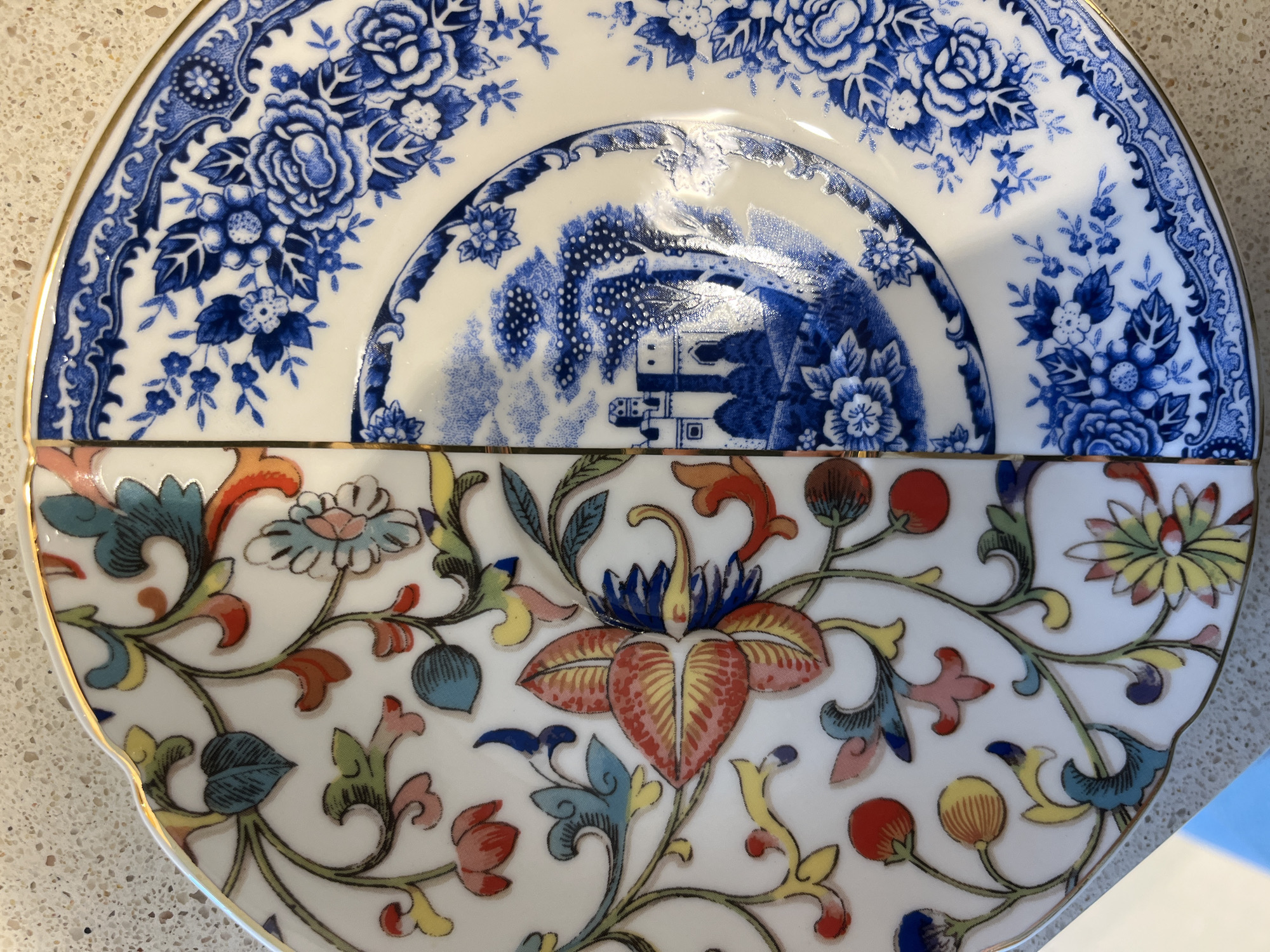 The image displays two ornate ceramic tableware pieces, likely plates or saucers, arranged vertically and partially overlapping. The scene is a still life.
The top piece is a white ceramic dish featuring an intricate blue and white pattern. Its outer edge showcases a dense floral and scrollwork border, with larger, stylized roses and smaller blossoms. Towards the center, a distinct "Willow Pattern"-like motif is visible, depicting traditional East Asian architecture, possibly pagodas or houses, surrounded by trees and landscape elements. The glazed surface shows some reflections from an overhead light source.
The bottom piece is a white ceramic dish with a vibrant, multicolored floral and foliate design. The pattern consists of stylized flowers and curling leaves in various shades of red, orange, yellow, green, and blue, all outlined in black. One prominent central flower exhibits red-orange petals with yellow accents and a blue center. This piece has a slightly scalloped edge adorned with a thin gold rim.
Both dishes are resting on a light-colored, speckled surface, resembling a countertop or table. No specific activity or event is taking place, and no text is visible on either piece of tableware or in the background. The items are located in Belgrade, Serbia.