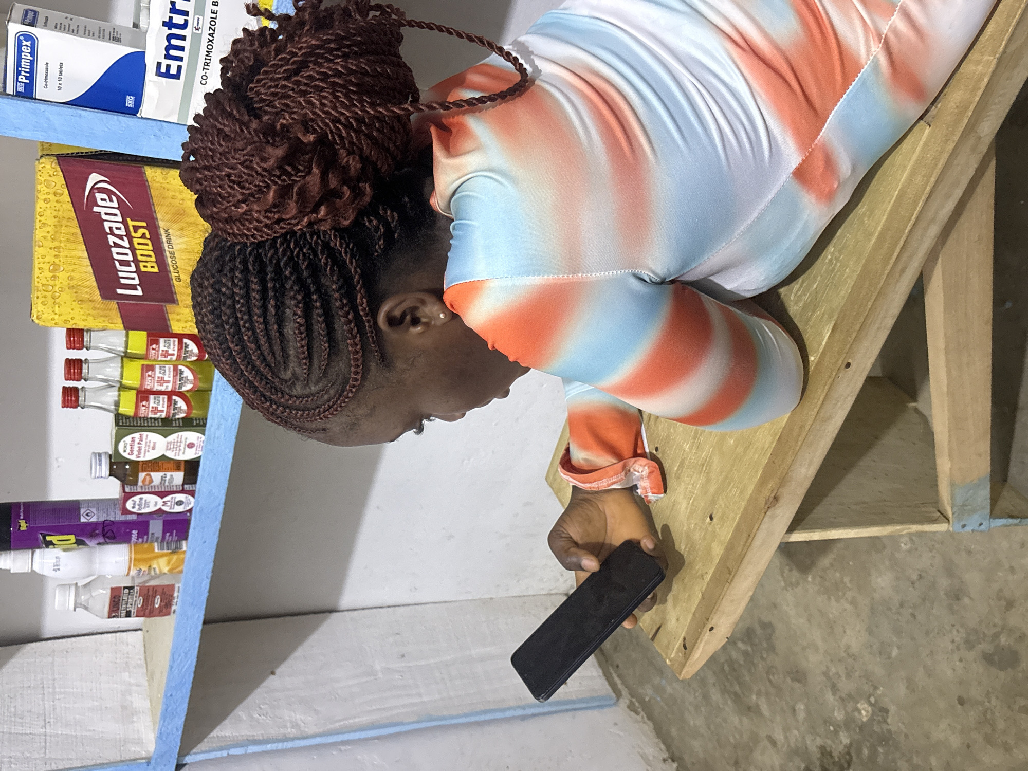 A woman with braided hair holds a smartphone. She leans on a wooden surface next to a shelf of pharmaceuticals. On the shelf are boxes of Primpex and Emtrim, a box of Lucozade Boost, bottles of iodine, and bottles of Gentian Violet Paint. The setting suggests a pharmacy or store in Ikot Ekpene, Nigeria.