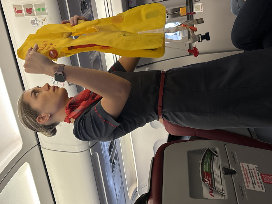 Inside a commercial aircraft cabin, a female flight attendant with blonde hair in a bun wears a dark grey short-sleeved uniform dress with red embroidery, a red neck scarf, and a red belt. A purple-banded smartwatch is visible on her left wrist. She holds a yellow life vest marked "DEMO ONLY" with both hands, presenting it. Her gaze is directed upwards and to the right. The background shows white overhead bins, various safety instruction decals (fire extinguisher, oxygen mask, life vest, green exit signs), and the top of a red-upholstered passenger seat. A safety information card, partially in Russian, is in the seatback pocket, and text below it includes "FASTEN SEAT BELT WHILE SEATED." The aircraft is situated in the city of Novogorelovo, Russia.