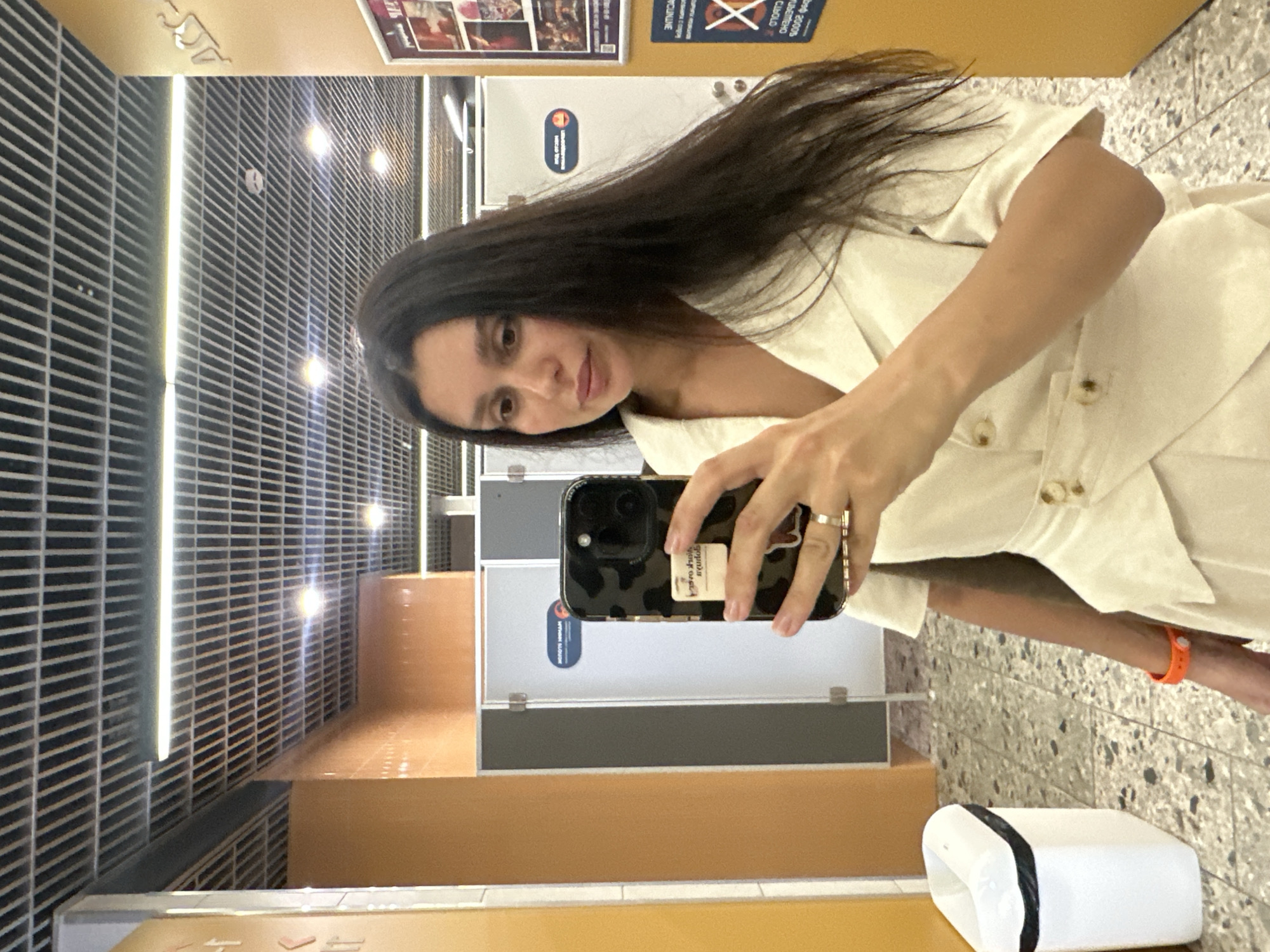 A young woman with long, dark hair takes a mirror selfie in what appears to be a public restroom or changing room, likely in Votkinsk, Russia, given the provided location. She is wearing a light-colored, button-down dress. Her left hand, adorned with a ring, holds up a smartphone with a patterned case, reflecting the scene. An orange bracelet is visible on her right wrist. The environment features tiled flooring, orange and gray walls, and a suspended ceiling with integrated lighting. A white trash can with a black liner is positioned to the left of the frame. Several doors, possibly for stalls, are visible in the background, along with signage. The lighting is bright, creating a slightly washed-out effect.