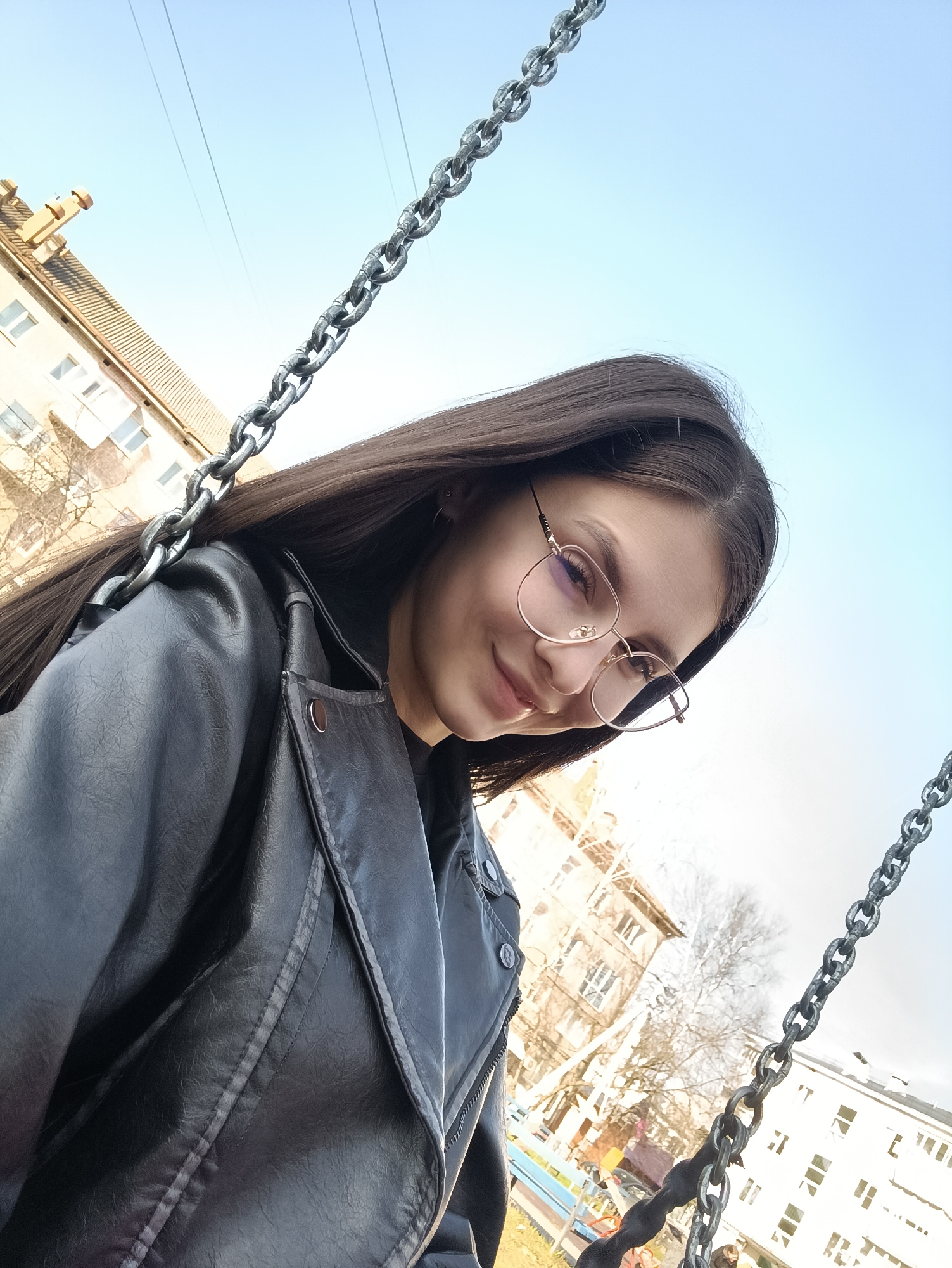 A young woman with long dark hair and wire-rimmed glasses is the focus of this selfie, taken from a low angle. She's wearing a black leather jacket. The background includes a clear blue sky and buildings, suggesting an urban environment. Chains of a swing set frame her face, indicating she's likely in a playground. The location is in the city of Unknown, Unknown.