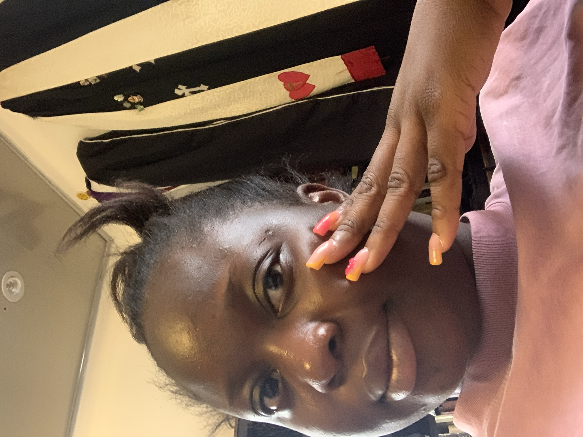 A medium shot captures a young Black woman from the shoulders up, in a selfie-style pose. She is looking towards the camera with her left hand resting near her face. The woman has dark skin and her hair is pulled up with a few strands loose around her face. She is wearing a light pink, high-necked top. Her fingernails are long and painted with a gradient of orange and pink. In the background, a black garment with a white stripe and decorative elements is visible, along with a light-colored wall. The lighting is bright, illuminating the woman's face and the details of the background.