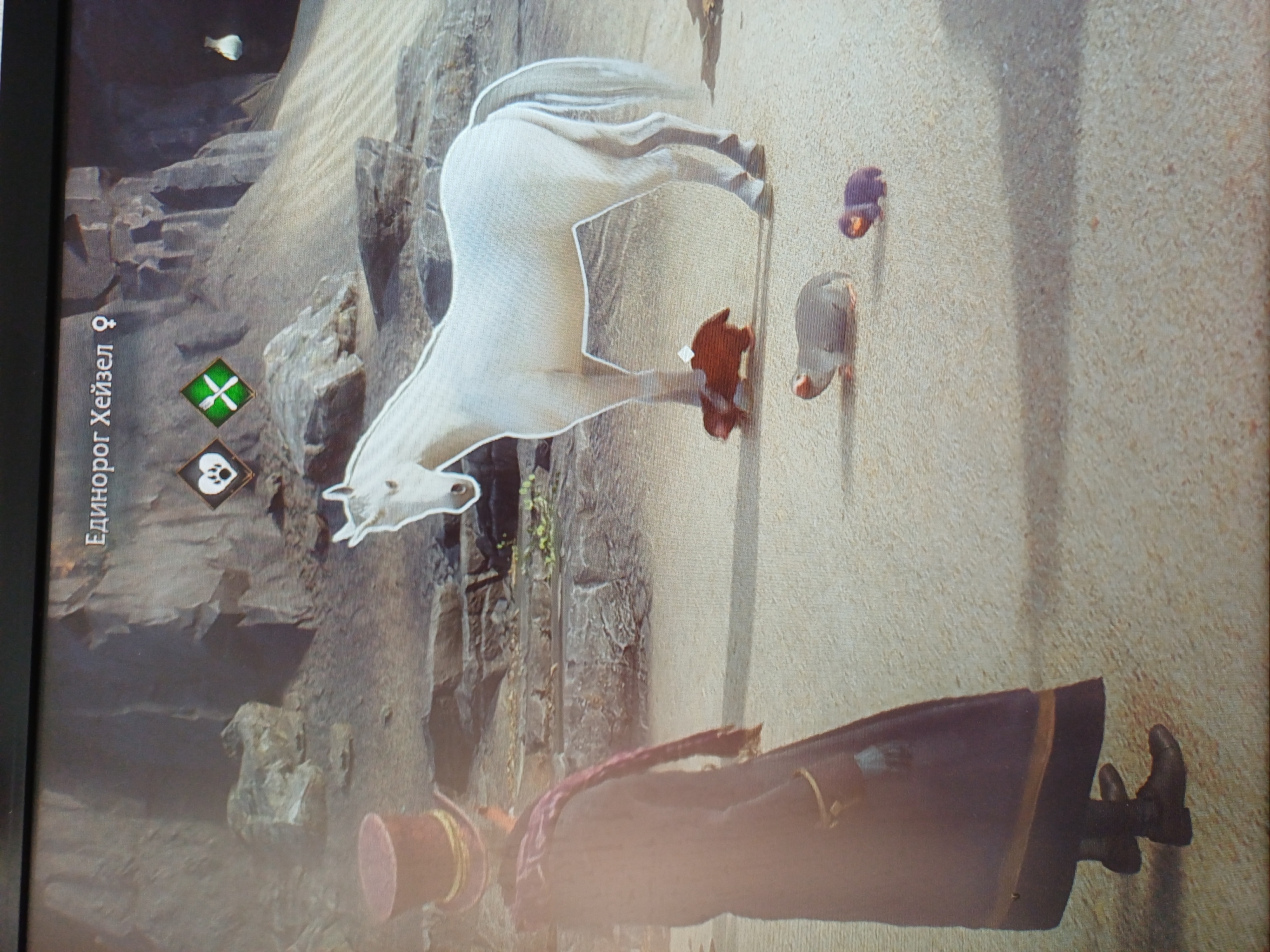The image shows a video game scene with a white horse, a person in a long dark coat and top hat, and three small creatures on a sandy, rocky terrain. The horse is the central focus, with the person standing to its left, facing away from the viewer. The small creatures include a brown one near the horse's legs, a grey and white spotted one, and a dark purple one. The environment appears to be a desert or canyon area with rock formations and sand. Text at the top of the screen indicates a creature named "Единорог Хейзел" and displays icons of a paw print inside a heart and a green diamond with a crossed knife and fork. The scene suggests the player is interacting with or observing these creatures within a game.