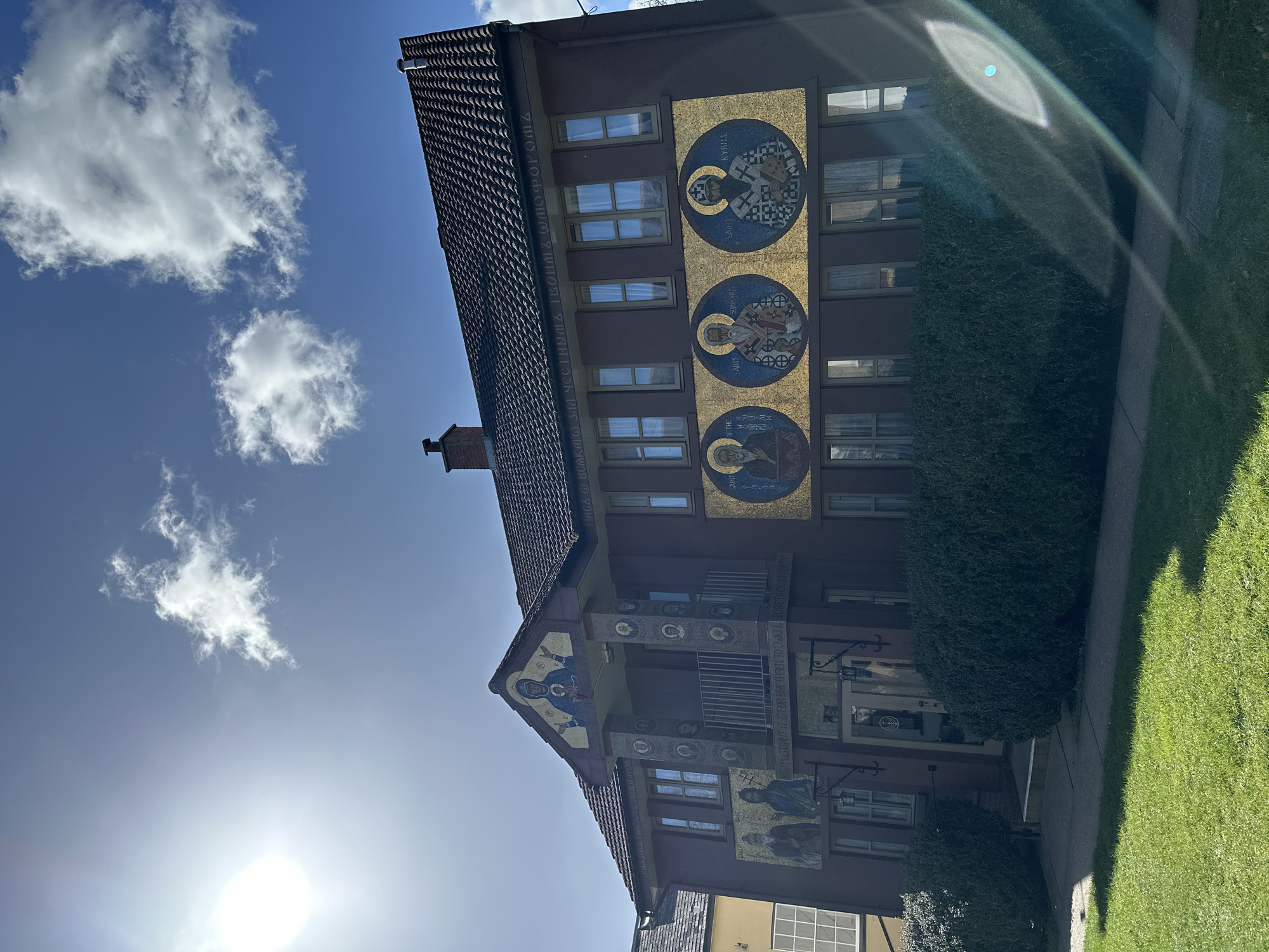 This image captures the exterior of a two-story religious building, identified as located in Maldon, United Kingdom, under a bright blue sky with scattered white clouds. The sun is visible in the upper left, causing a distinct lens flare across the lower right portion of the frame.
The building's main facade is dark maroon, featuring a brown tiled roof with a brick chimney on the left and a metal vent on the right. A prominent pediment or gable at the center of the roofline houses a mosaic depicting the Virgin Mary holding the Christ Child, set against a gold background, with arms outstretched. Below this, a recessed balcony area with a metal railing is supported by two square pillars. These pillars are adorned with multiple circular mosaic portraits of various figures. Below the balcony, to the left, are two full-figure mosaic portraits of bearded male saints. An inscription in Cyrillic script runs along the frieze below the roofline, and another English inscription, "HOW PLEASANT IT IS FOR BRETHREN TO DWELL TOGETHER IN UNITY," is visible below the balcony railing.
To the right of the balcony, three large circular mosaics are displayed against a gold tessellated background. Each mosaic depicts a bearded male figure in religious vestments. Partially legible labels identify them as "SAINT OF THE...", "SAINT SAHROLES," and "SAINT KYRILL." The building features numerous rectangular windows with light-colored frames on both levels.
The ground level presents a double-door entrance with glass panels, flanked by two wall-mounted black lanterns. A paved pathway leads to this entrance. The immediate foreground consists of a neatly trimmed green lawn, separated from the building by a dense, dark green hedge that runs along the facade. To the far left, a portion of an adjacent lighter-colored building with white-framed windows is visible.