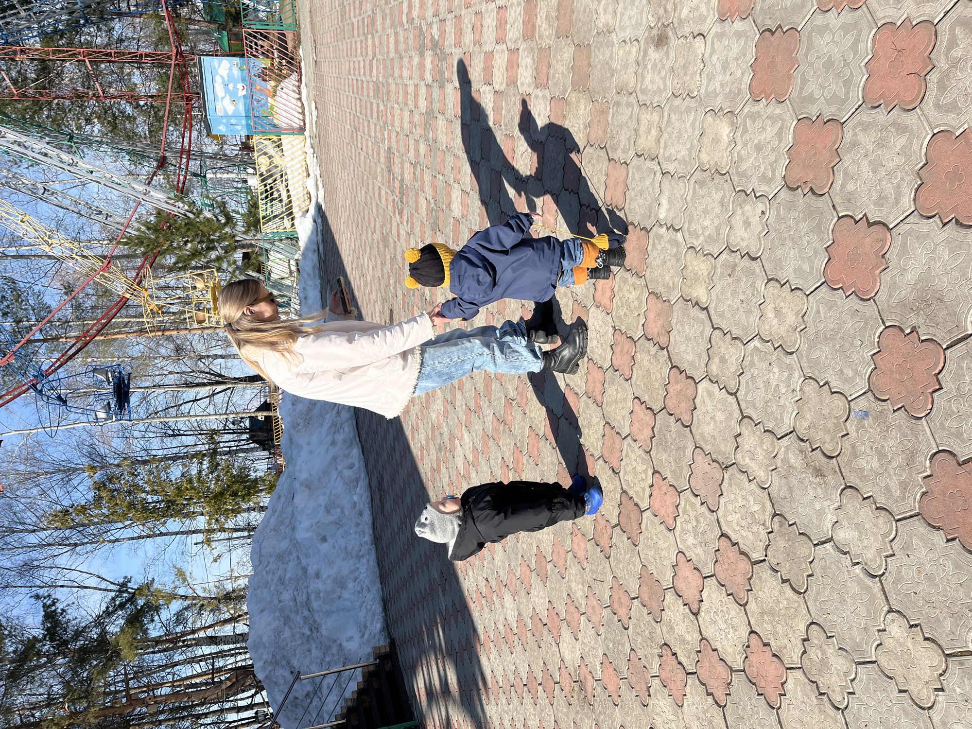 A woman and two children are on a paved area in Chaikovsky, Russia, near an amusement park on a sunny day. The woman, with long blonde hair, is wearing a light-colored jacket and jeans. She is holding the hand of a toddler, who is wearing a blue jacket, jeans, a black hat with yellow pom-poms, and orange boots. A second child, wearing a dark jacket, gray hat, and blue shoes, is lying on the ground. The ground is paved with patterned tiles. A snow bank is visible on the left side of the image, and trees and a Ferris wheel are in the background. Shadows are cast on the pavement by the figures.