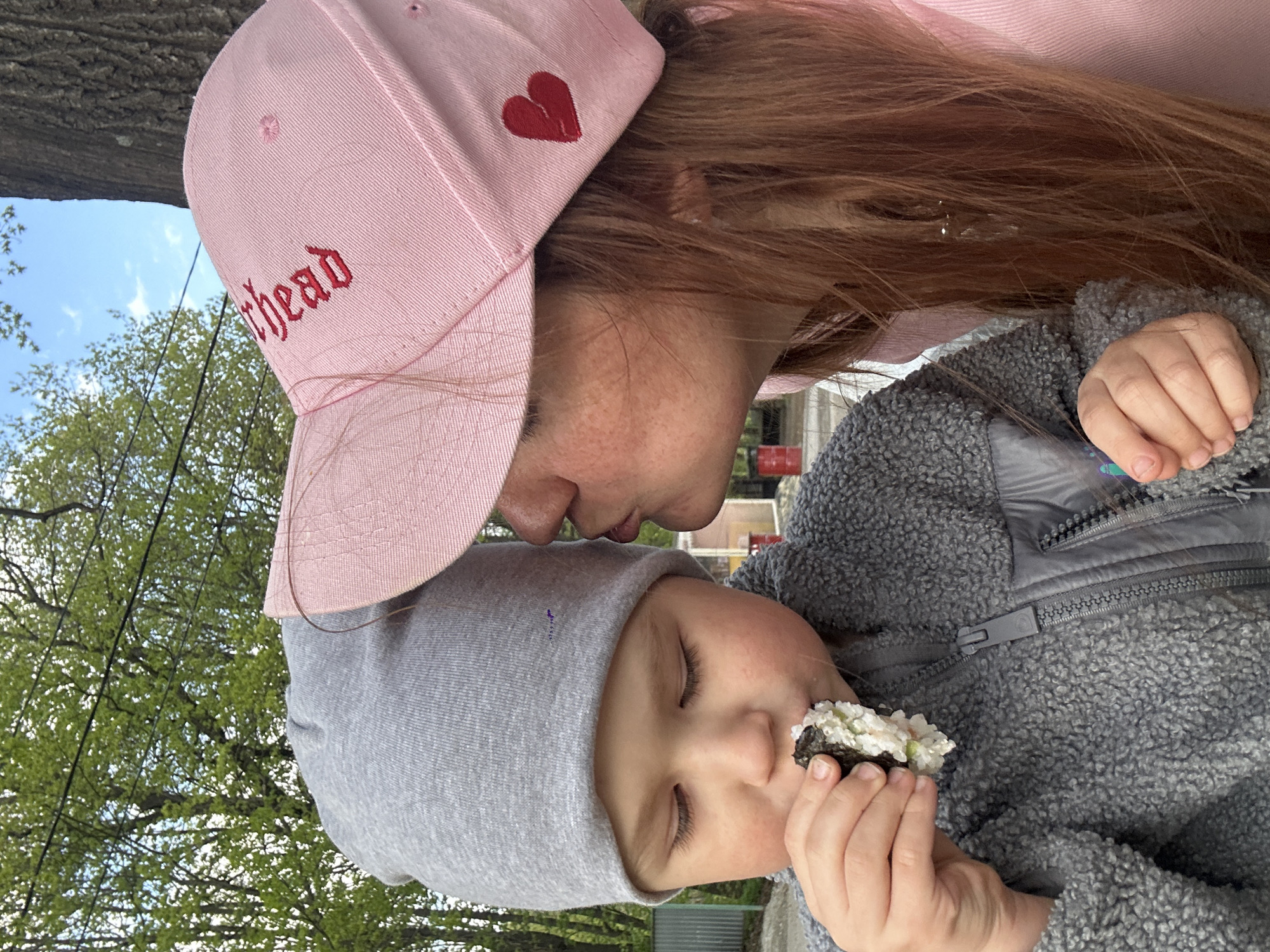 A woman and a young child are outdoors. The woman wears a pink baseball cap with a red heart and the word "thead" embroidered on it, and a pink top. She has red hair and freckles. The child wears a gray hat and a gray fleece jacket. The child is eating a sushi roll. The scene is in a park, with trees and blue sky visible in the background. Based on the style of architecture and vegetation, the location may be in a city like Nizhny Novgorod, Russia.
