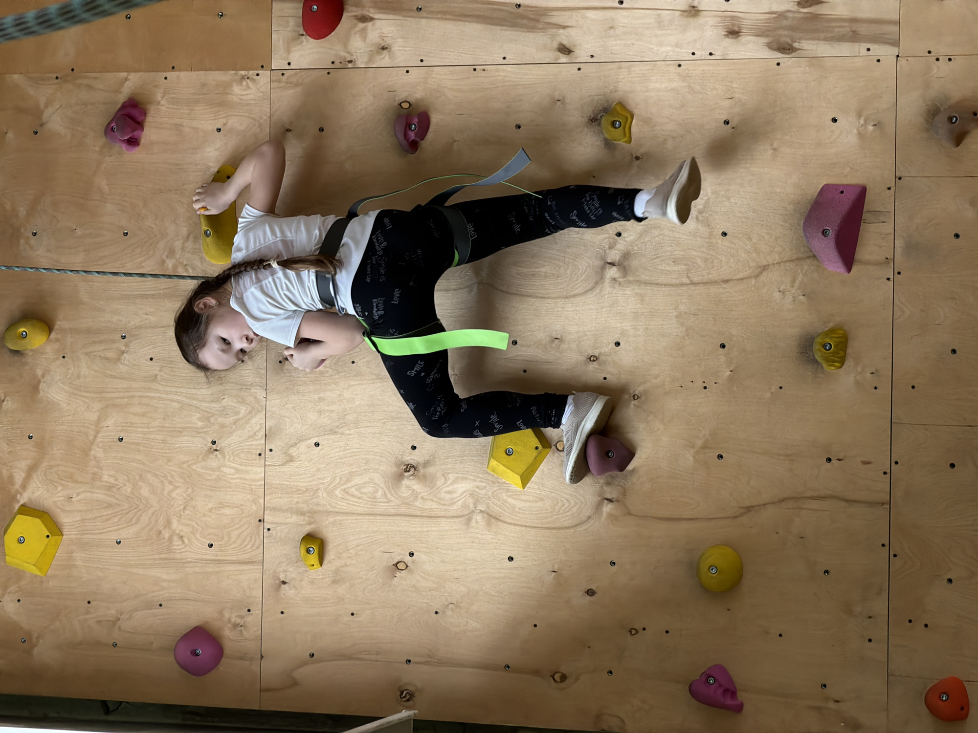 A young girl is depicted mid-activity on an indoor climbing wall in Izhevsk, Russia. She is wearing a white t-shirt, black leggings patterned with text, and light-colored sneakers, secured by a safety harness with gray and neon green straps. The climbing wall is made of light-toned wooden panels and features numerous colored climbing holds in shades of red, pink, yellow, and purple. A climbing rope is visible, extending from the upper left. The girl is positioned horizontally across the wall, looking towards the viewer with a neutral, focused expression. Her right hand grasps a yellow hold, and her left foot is placed on a purple hold. Visible text on her leggings includes words like "Love", "Smile", "Kindness", "Make Up", and "Positive", repeated multiple times. The setting suggests an indoor recreational climbing facility.