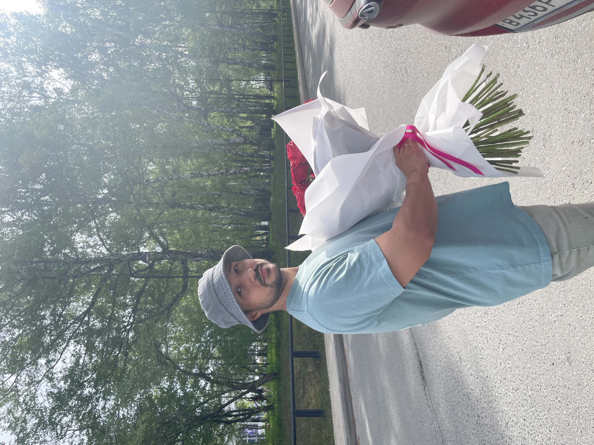 A man wearing a light blue t-shirt and a grey bucket hat holds a bouquet of red roses wrapped in white paper tied with a pink ribbon. He stands on a grey asphalt road next to a maroon-colored vehicle, the license plate of which is partially visible with the characters "B456P". Behind the man, there is a row of trees, and a green metal fence separates the road from a grassy area. The scene is outdoors, and based on the trees, it appears to be a park area. According to the information provided, the location is in the city of Izhevsk, Russia.
