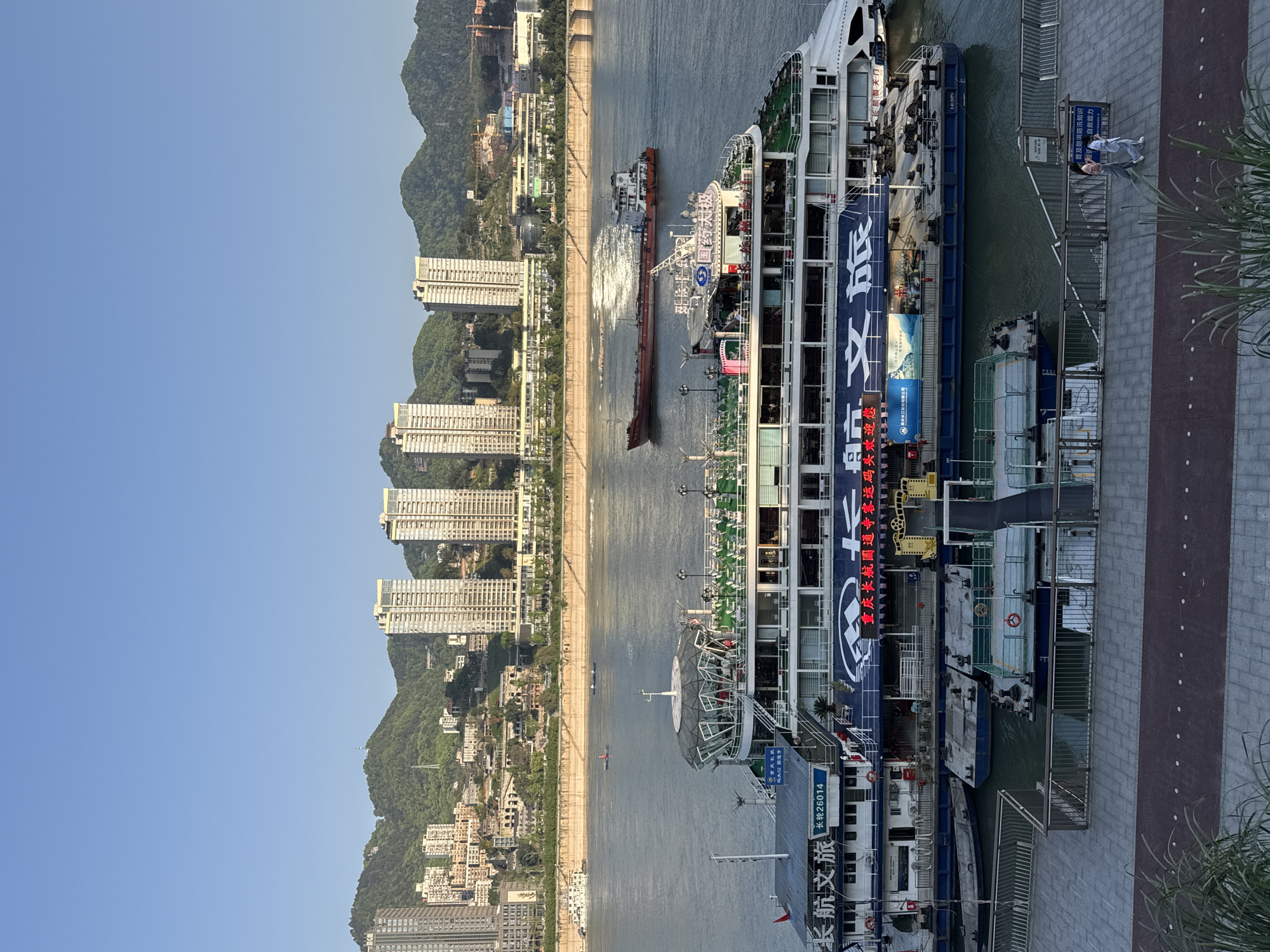 This aerial landscape depicts docked cruise ships with a city on the opposite bank. The city skyline is dominated by high-rise residential buildings backed by mountain terrain under a clear blue sky.
On the right side of the frame, a stone-paved walkway borders the dock where the cruise ships are moored. Signage in Chinese is visible, providing information and safety guidelines. People are present on the walkway near the boats. A cruise ship is labeled "Changhang Wenlv Changlun 26014". Additional text on the cruise ship indicates the location is a passenger transportation terminal, welcoming visitors.
A second ship is seen on the river. Another boat is creating a noticeable wake behind it, indicating it is in motion.
