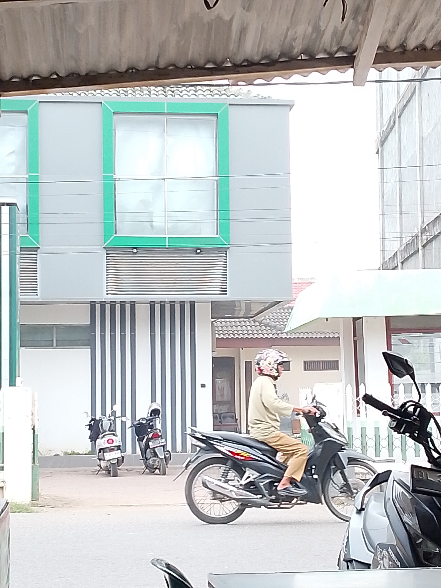 A medium shot captures a street scene in Lhokseumawe, Indonesia, featuring a man riding a black motorcycle. The man, wearing a helmet and a light brown long-sleeved shirt and tan pants, is in motion. To his left, two white scooters are parked. In the background, a building with a gray facade and green trim is visible, along with more scooters and building structures. The scene appears to be during daylight.
