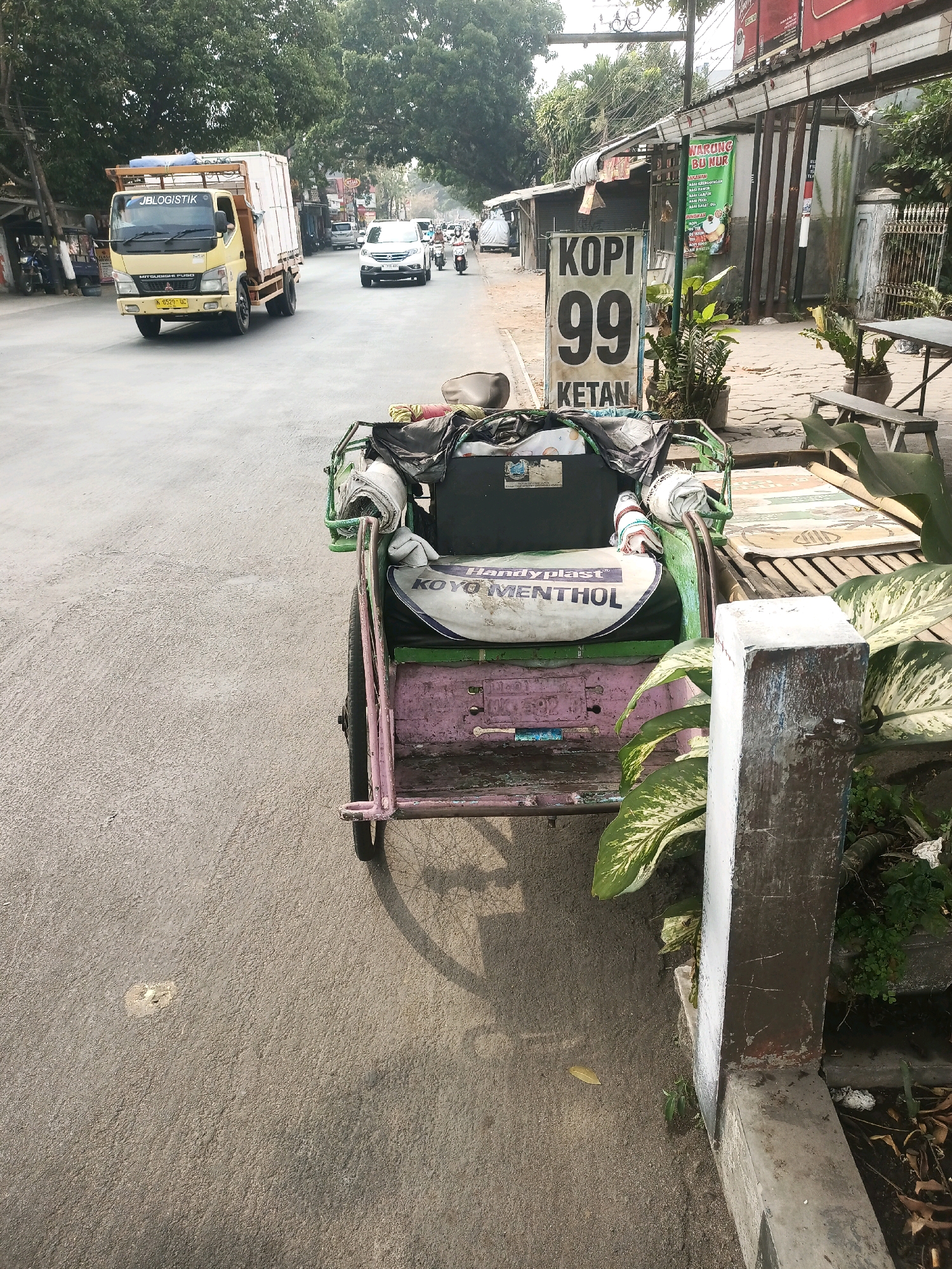 In the city of Malang, Indonesia, a road scene unfolds with a yellow delivery truck, labeled "JBLOGISTIK" and a Mitsubishi FUSO model, traveling on the asphalt. A white car follows behind it, also on the road. To the right of the road, a pedestrian walkway is visible, featuring a sign that reads "KOPI 99 KETAN." Beside the sign, a small, weathered pink and green rickshaw is parked, laden with assorted items wrapped in cloth and plastic. The rickshaw is positioned next to a concrete post and lush green plants. Further back, a building with a corrugated metal roof appears to house shops or stalls, and trees line the street, casting shadows on the scene.
