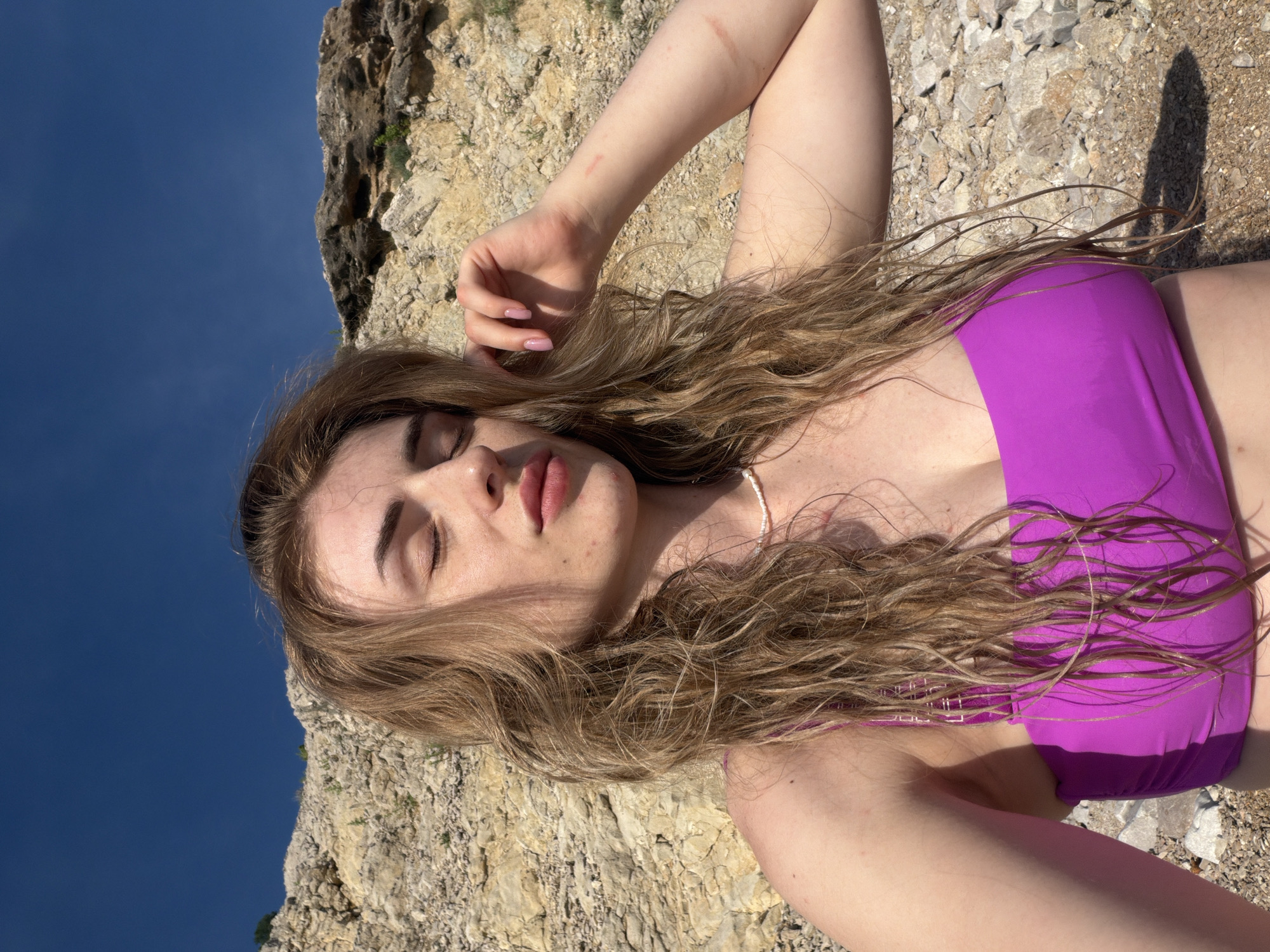 A woman with long, wavy blonde hair wears a purple bikini top and a pearl necklace. She is taking a selfie outdoors, with her eyes closed and her hand raised to her forehead. Behind her, a rocky cliff face meets a deep blue sky. The ground is covered in pebbles and rocks. The woman has several small scratches on her right arm. The scene appears to be a beach or coastal area.