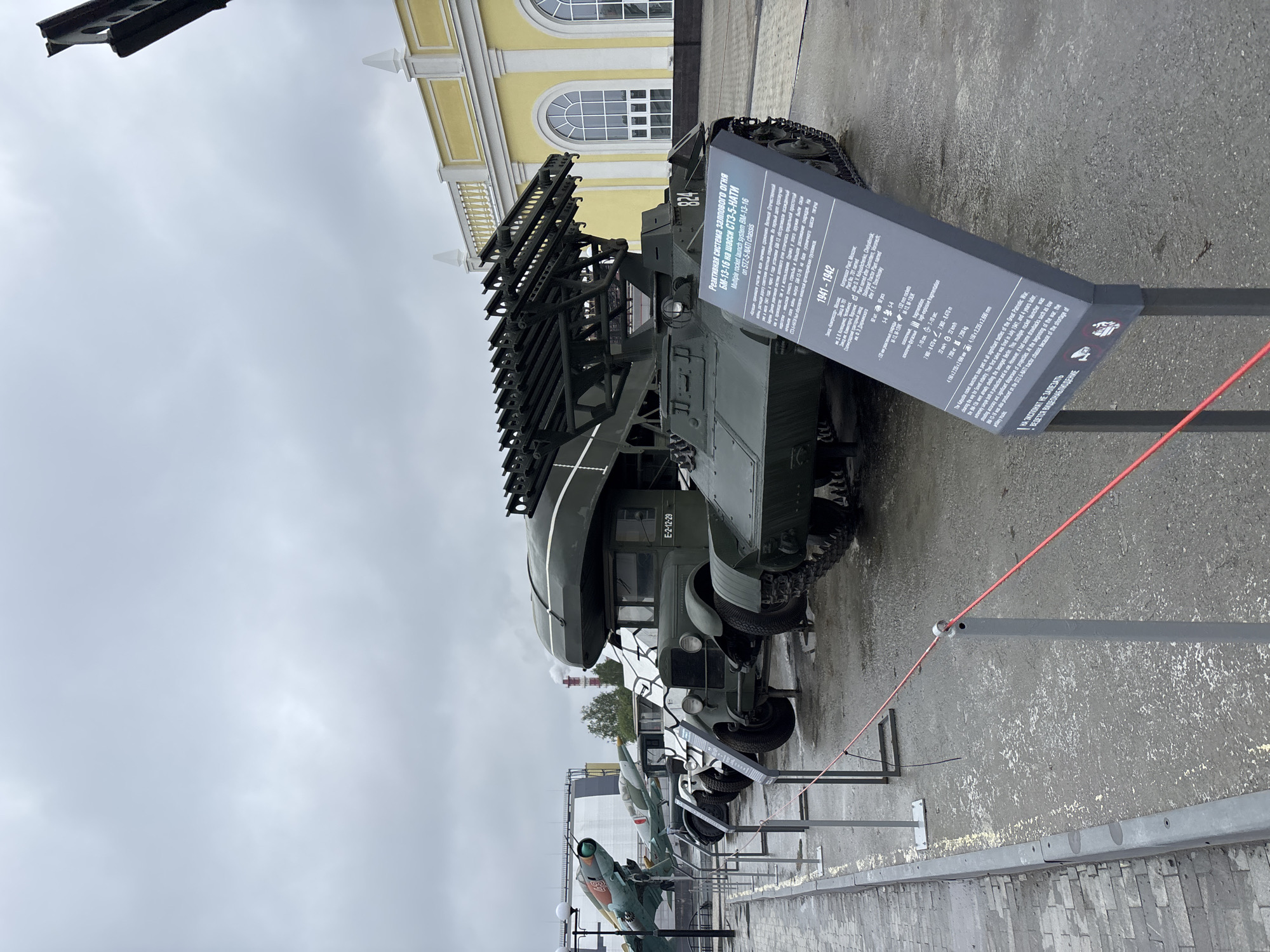 In Verkhnyaya Pyshma, Russia, an outdoor exhibition of military equipment is displayed under an overcast sky. The foreground features a row of several military vehicles, including a rocket launcher system mounted on a tracked chassis, identified as BM-13-16 on a GAZ-51 chassis. Next to it is a vintage truck with the license plate E-2-12-29. Further to the left, several fighter jets are suspended on stands. A bright yellow building with arched windows stands in the background. A descriptive plaque for the BM-13-16 rocket launcher is visible in front of the vehicle, providing specifications and historical context. Red ropes on metal posts cordon off the exhibits, indicating a museum or exhibition ground. The ground is wet, suggesting recent rain. No people are visible in the scene.