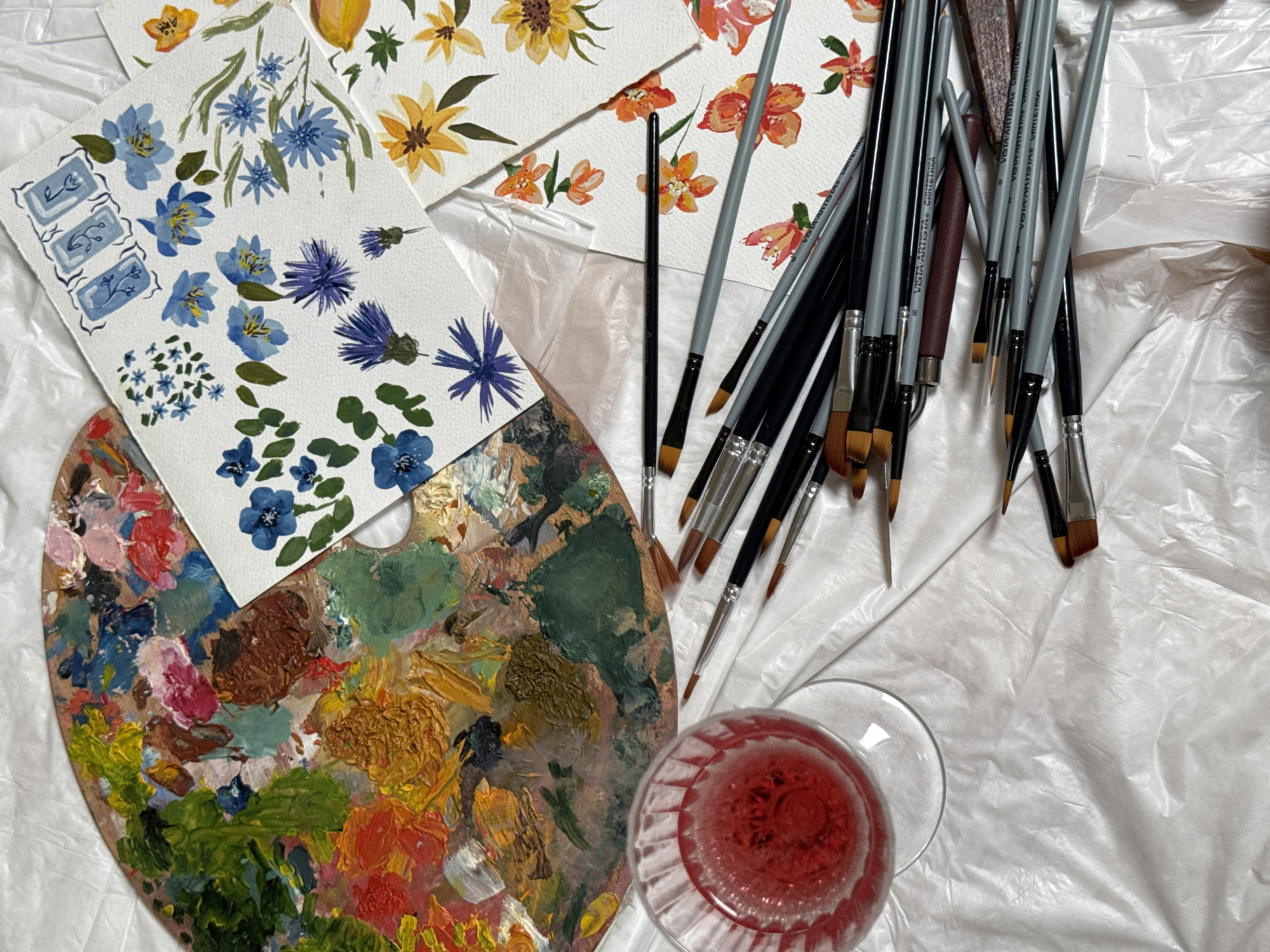 The image shows an artist's workspace. There is a wooden palette with various globs of colorful paint, including red, green, yellow, brown, and blue. Several watercolor paintings of flowers are scattered around, featuring blue, purple, yellow, and orange blooms. A collection of paintbrushes with grey and black handles lies near the paintings. A glass of red liquid sits on the right side of the image. All items are arranged on a wrinkled white surface. There is no evidence in the photo to indicate that the scene is in Sapronovo, Russia.