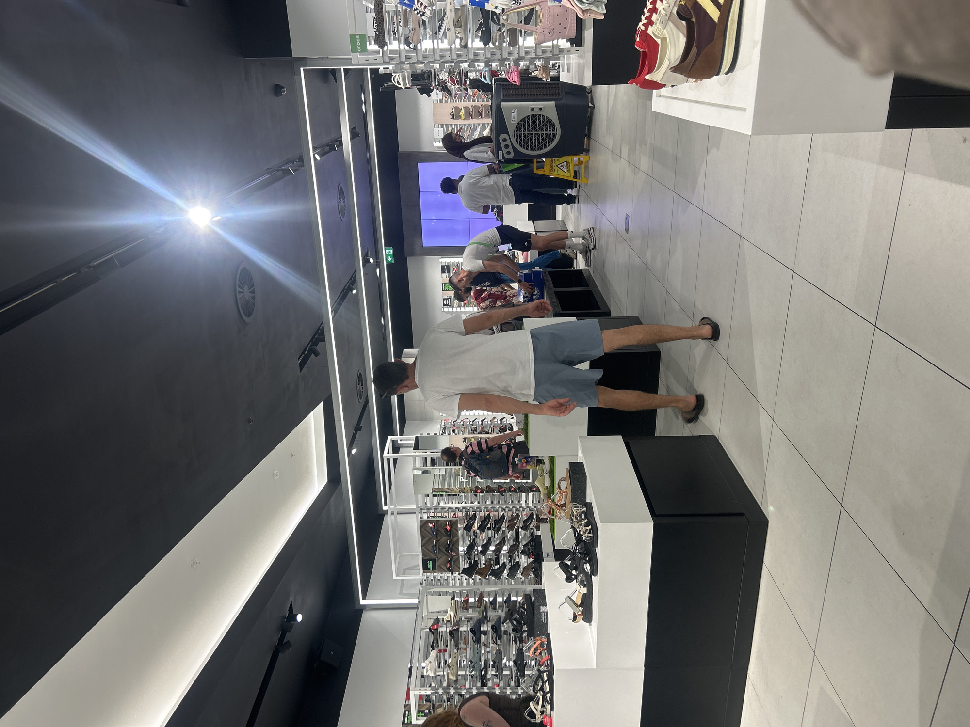 The image shows the interior of a shoe store in London, United Kingdom. The store is modern with white and black display shelves holding various shoe models. People are browsing, with one man in a white t-shirt and blue shorts standing in the foreground. Other customers are near the back, some looking at shoes and others near what appears to be an air conditioning unit. The lighting is bright, with prominent ceiling light fixtures. The floor is tiled in a light color, and there are displays of shoes along the walls.