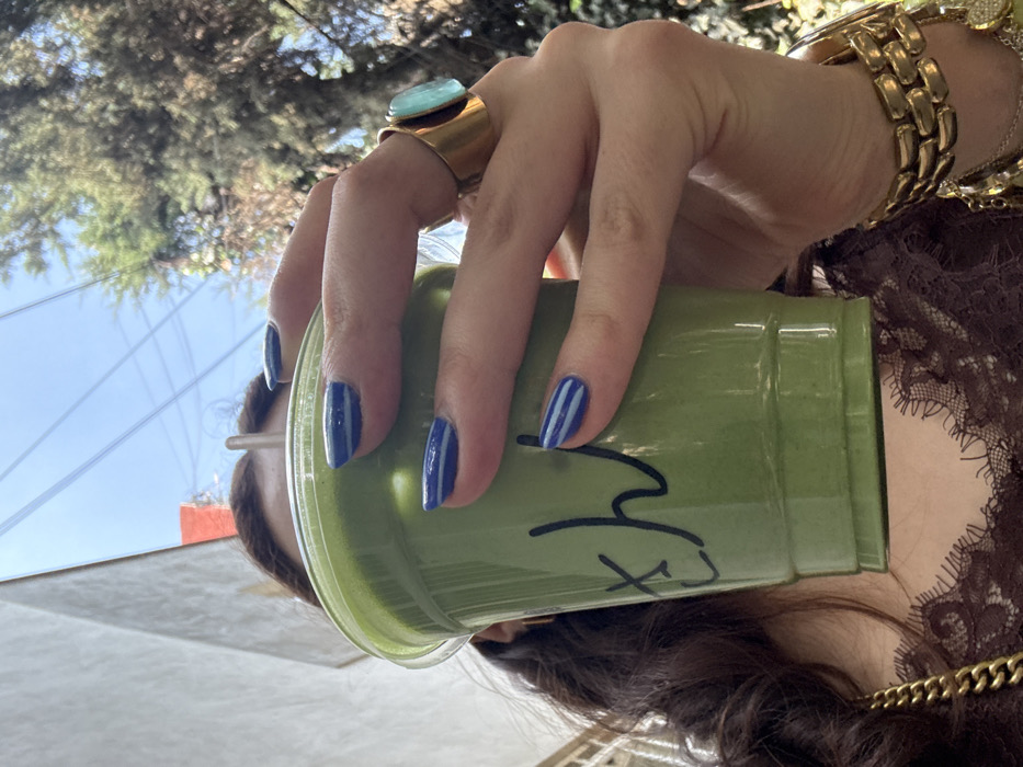 A close-up, low-angle shot captures a person holding a translucent green plastic cup containing a green beverage, with a straw visible. The person's right hand, prominently featured, has nails painted with blue polish, some accented with two thin white horizontal stripes. A large gold ring with an oval turquoise-colored stone is worn on the ring finger, and multiple gold chain bracelets are visible on the wrist. Black Japanese hiragana characters, "さん" (san), are written on the side of the cup. Partial view of the subject includes dark hair and a brown lace garment with a scalloped neckline. The background consists of a light-colored building wall to the left, vibrant green tree foliage to the right, and a clear blue sky overhead with faint utility lines. The scene is set in an outdoor urban environment within Mexico City, Mexico, likely in a neighborhood known for its cafes and tree-lined streets, such as Colonia Roma Norte or Condesa.