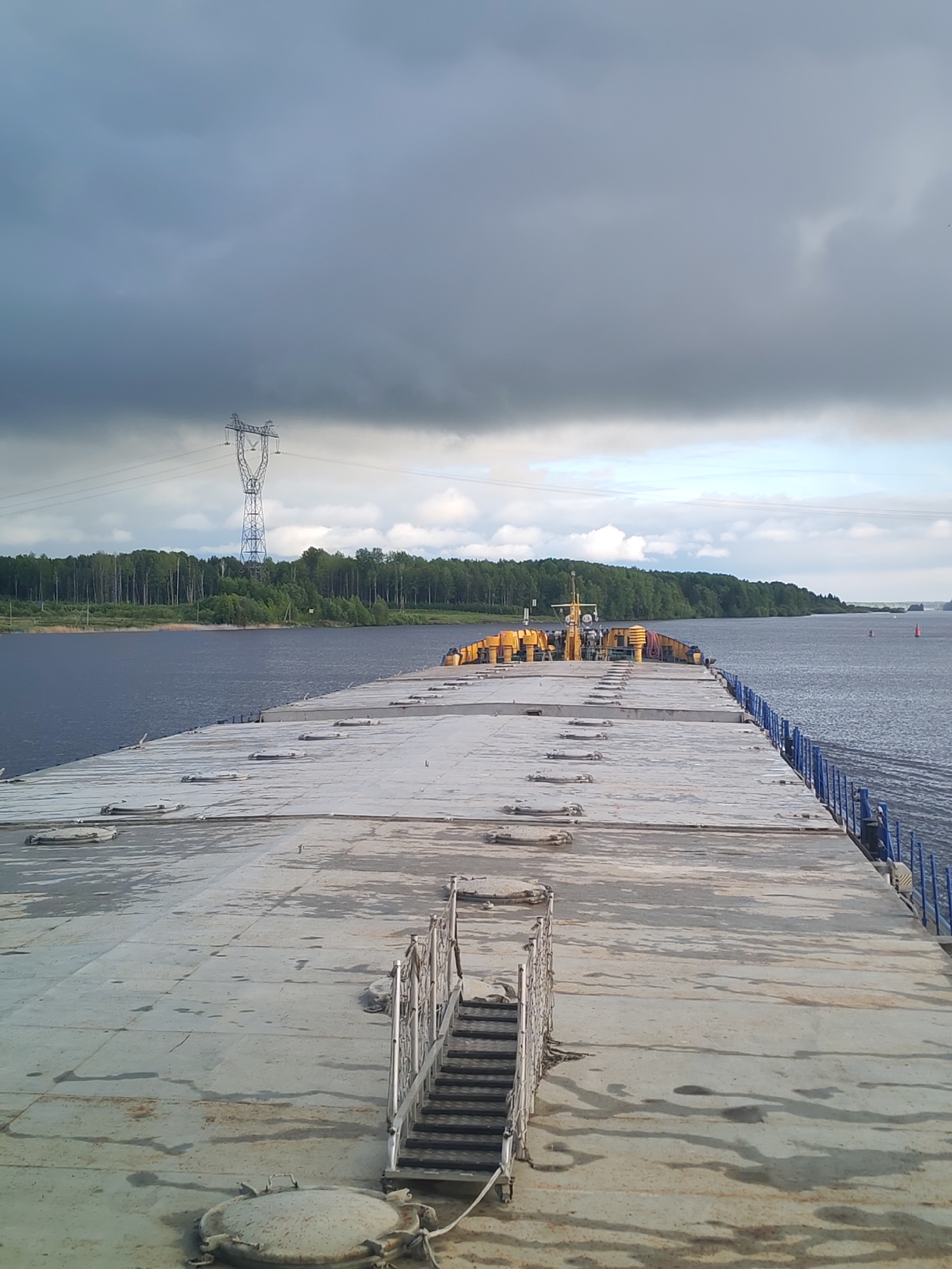 The image depicts the deck of a barge or flat-topped ship, likely on a river or wide body of water. The deck is concrete or a similar material, marked with seams and access hatches. A set of metal stairs is visible near the foreground. The railing along the ship's edge is blue. In the background, a line of trees runs along the shore of the water body. Further back is an electrical transmission tower. The sky is overcast with dark storm clouds dominating the upper portion of the image. No people are readily visible in the scene. The location is unknown.
