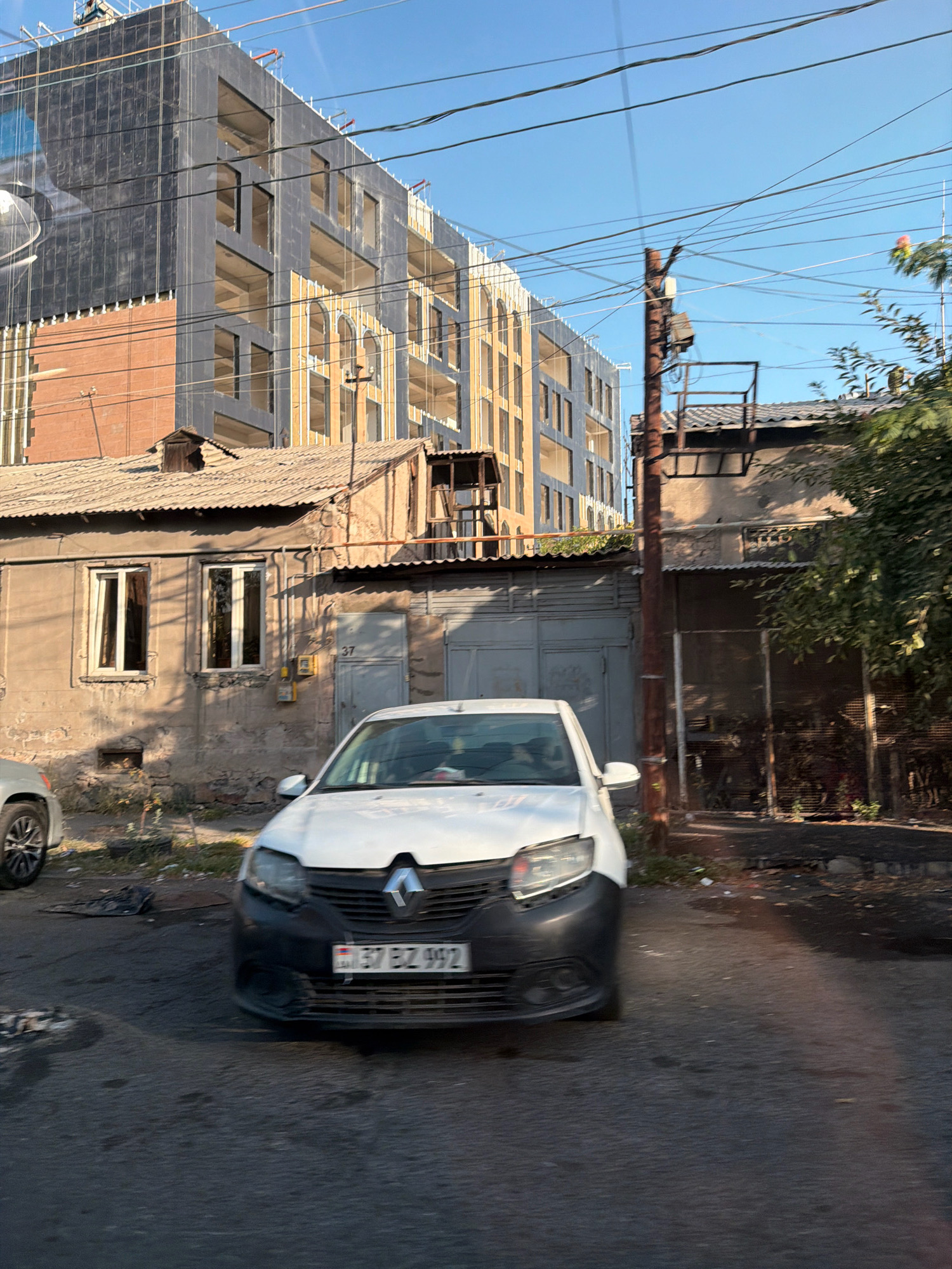 The image depicts a street scene in Yerevan, Armenia, on a clear day. In the foreground, a white Renault sedan with license plate "57 BZ 992" is parked, partially obscuring an older, single-story building with a corrugated metal roof. This building features two windows and a gray garage door with the number "37" displayed above it. Behind this older structure, a new, multi-story building is under construction, characterized by its unfinished facade with exposed concrete and dark paneling. Electrical wires crisscross the sky, typical of urban infrastructure. The ground is paved, showing some wear and tear. The overall impression is one of urban development, with a contrast between older and newer architecture.