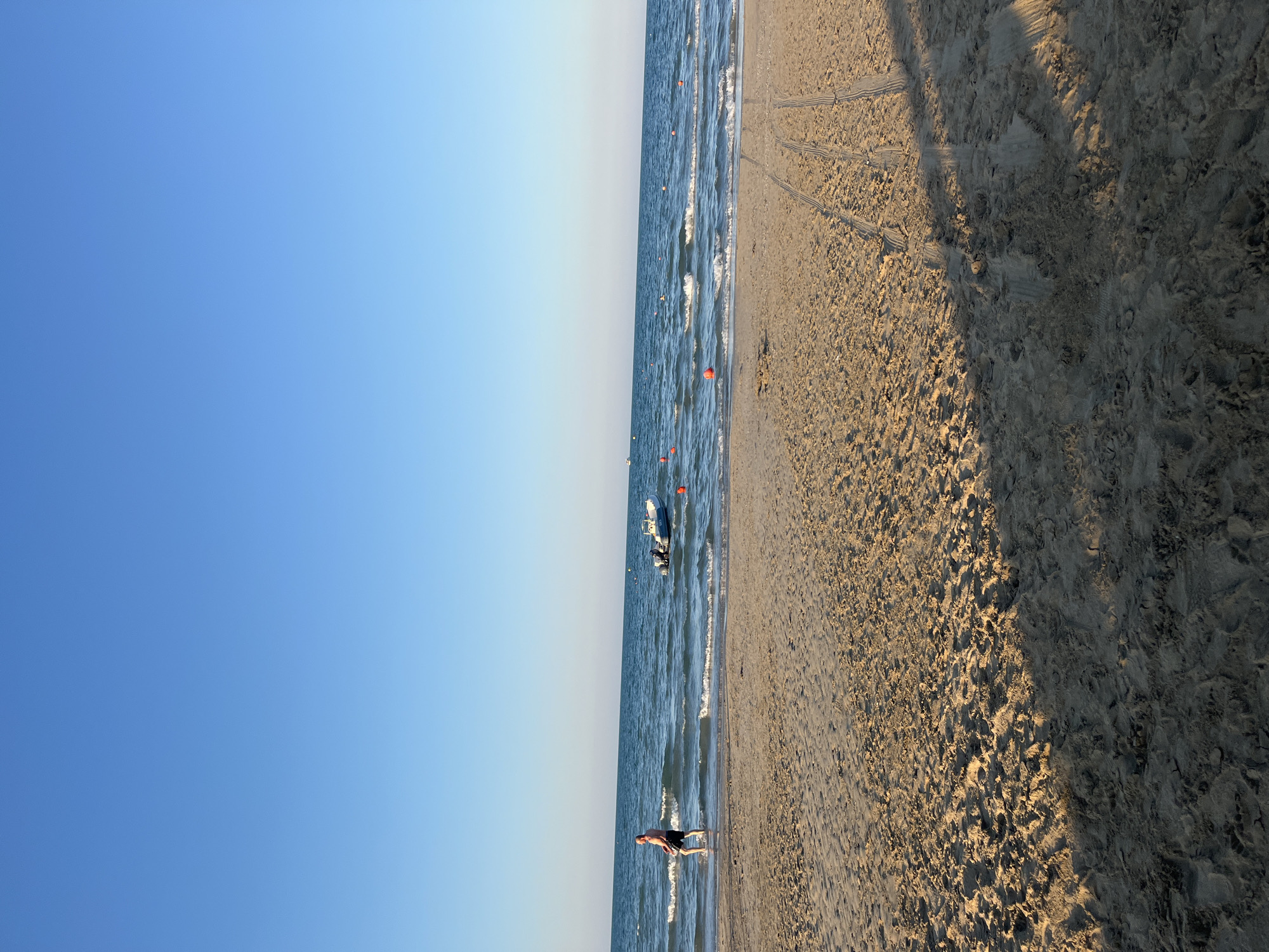 A man is walking in the shallow water at the edge of a sandy beach on a clear, sunny day. The sea is calm with small waves. In the distance, there is a small boat and several orange buoys. Tire tracks are visible in the sand in the foreground. This scene is consistent with the beaches of Rimini, Italy, known for their long stretches of sand and calm Adriatic Sea.