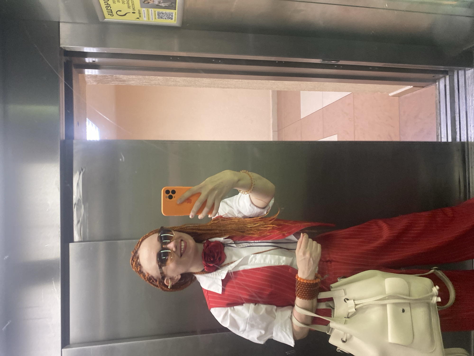 In an elevator, a woman with orange braided hair and sunglasses is taking a selfie. She is wearing a white shirt, a red pinstriped vest, and matching red pinstriped pants. She has a red fabric flower pinned to her collar and is accessorized with multiple bracelets. In her left hand, she holds an orange smartphone, and with her right hand, she carries a light-colored drawstring bag. The elevator doors are partially open, revealing a hallway with light-colored walls and tiled flooring. A yellow flyer with Cyrillic text and a QR code is visible on the right elevator door. The overall scene suggests the woman is in a residential building in Sochi, Russia.