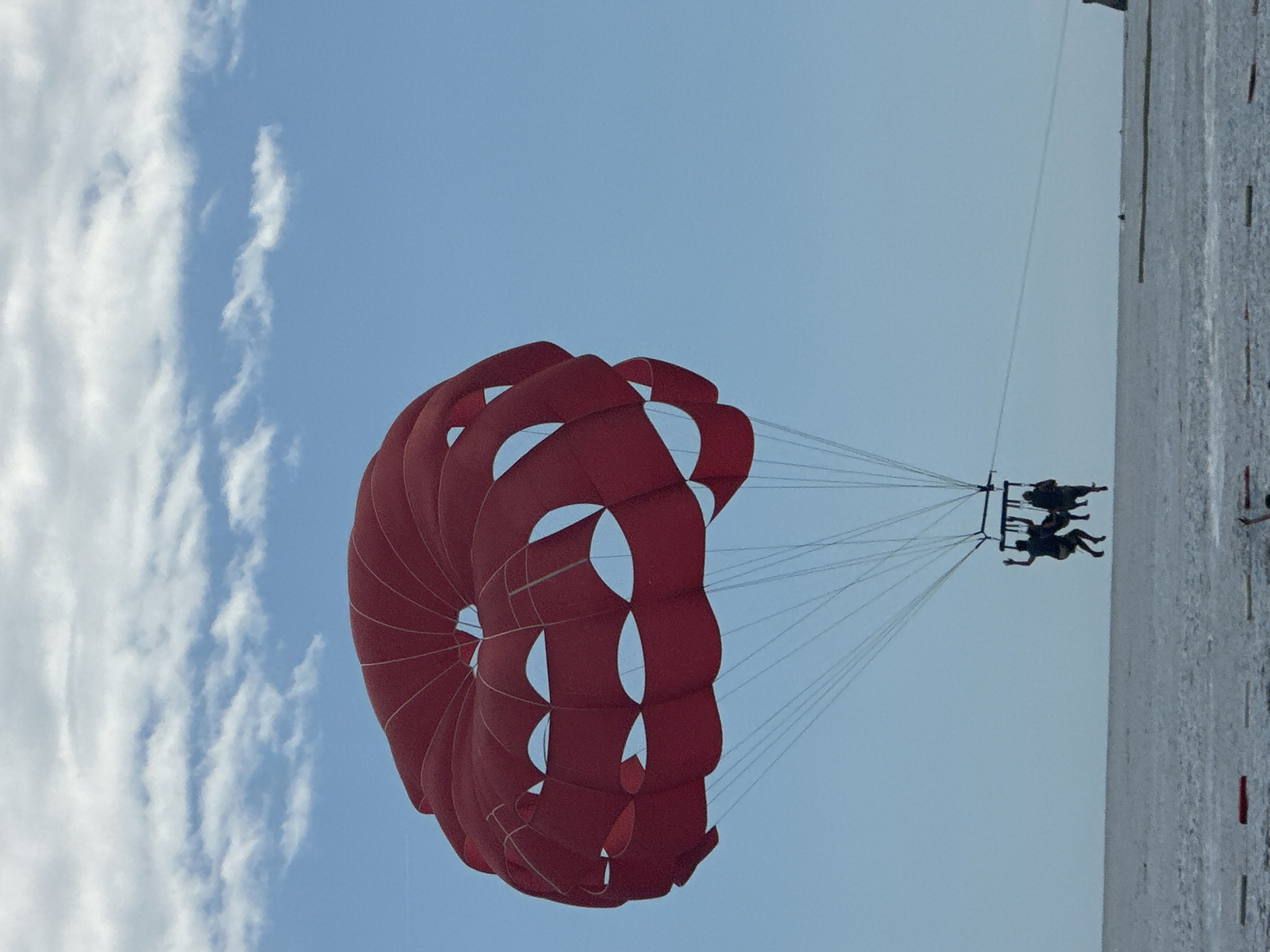 A photograph captures a vibrant scene of parasailing above the sea under a bright sky. A large, prominent red parasail, with its distinctive segmented design, is suspended high in the air, dominating the upper left portion of the frame. Below the parasail, two individuals are visible in a harness, silhouetted against the lighter sky as they are lifted by the activity. They are connected to the parasail by numerous thin ropes.
The sky is a clear, light blue with scattered white, wispy clouds visible on the left side of the image, indicating fair weather conditions ideal for such an activity. Below the parasailers, a calm expanse of blue-gray sea or ocean stretches across the bottom and right side of the frame, reflecting the brightness of the sky. The overall impression is one of leisure, adventure, and open air. This activity is taking place likely off the coast of Budva, Montenegro. There is no visible text in the image.