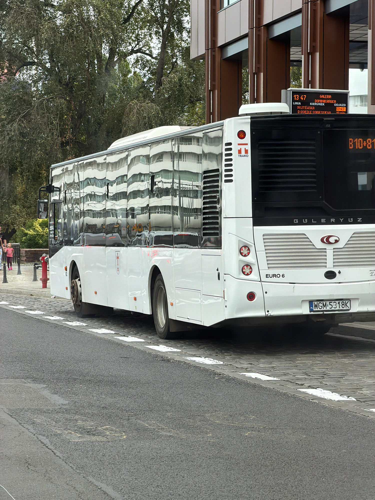 The image shows a white bus with the license plate "WGM 5318K" parked on a cobblestone street in Wrocław, Poland. The bus has a digital display on the back showing "B10+81". The bus is reflecting the surrounding environment, including trees and buildings. To the left of the bus, a red fire hydrant is visible. In the background, two women are walking on the sidewalk. The scene appears to be a typical urban street in a city.