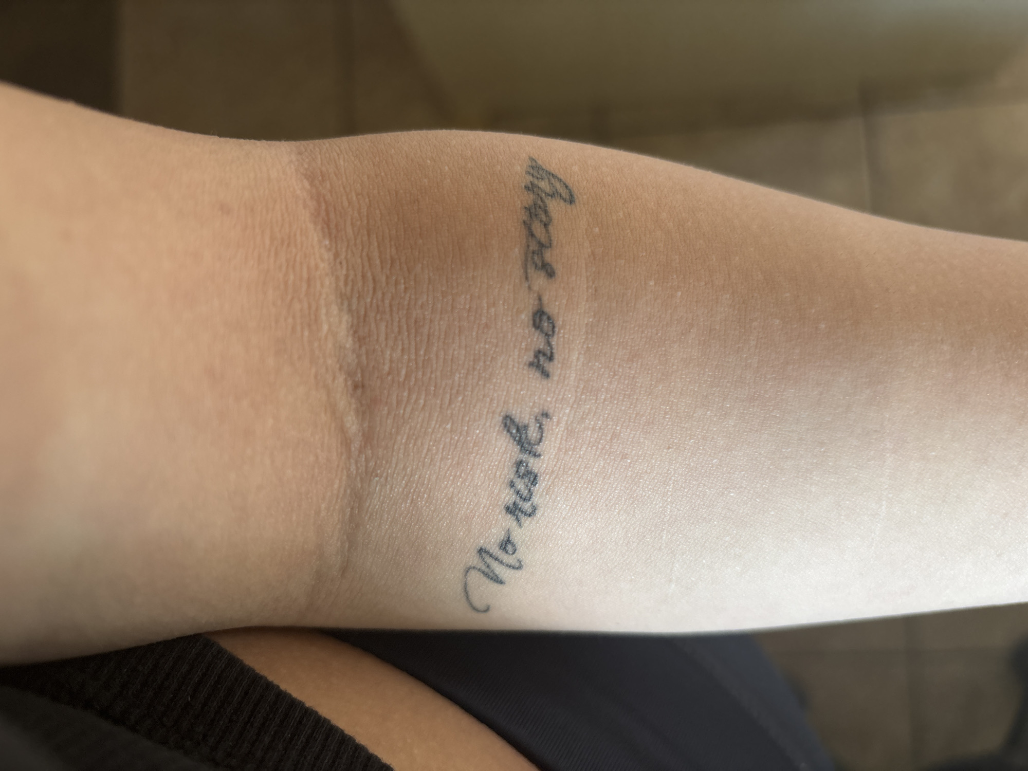 This close-up image, captured in Orizari, Bulgaria, features the inner arm of a person. The arm extends diagonally across the frame, displaying light to medium-toned skin with visible texture, particularly around the creased area of the inner elbow, indicating the arm is bent. A dark-colored, possibly ribbed, fabric from an article of clothing is partially visible along the bottom edge of the frame.
The main subject of the image is a tattoo on the inner forearm/bicep. Written in black cursive script, the tattoo is oriented vertically along the arm and clearly spells out the phrase: "No risk, no story." The lettering appears distinct and well-preserved. The background is blurred and indistinct, showing neutral brown and beige tones, suggesting an indoor setting. No specific activities or emotions are discernible, and no other prominent objects or detailed location cues are visible beyond the arm and its tattoo. The image focuses on this anatomical detail and its inscription.