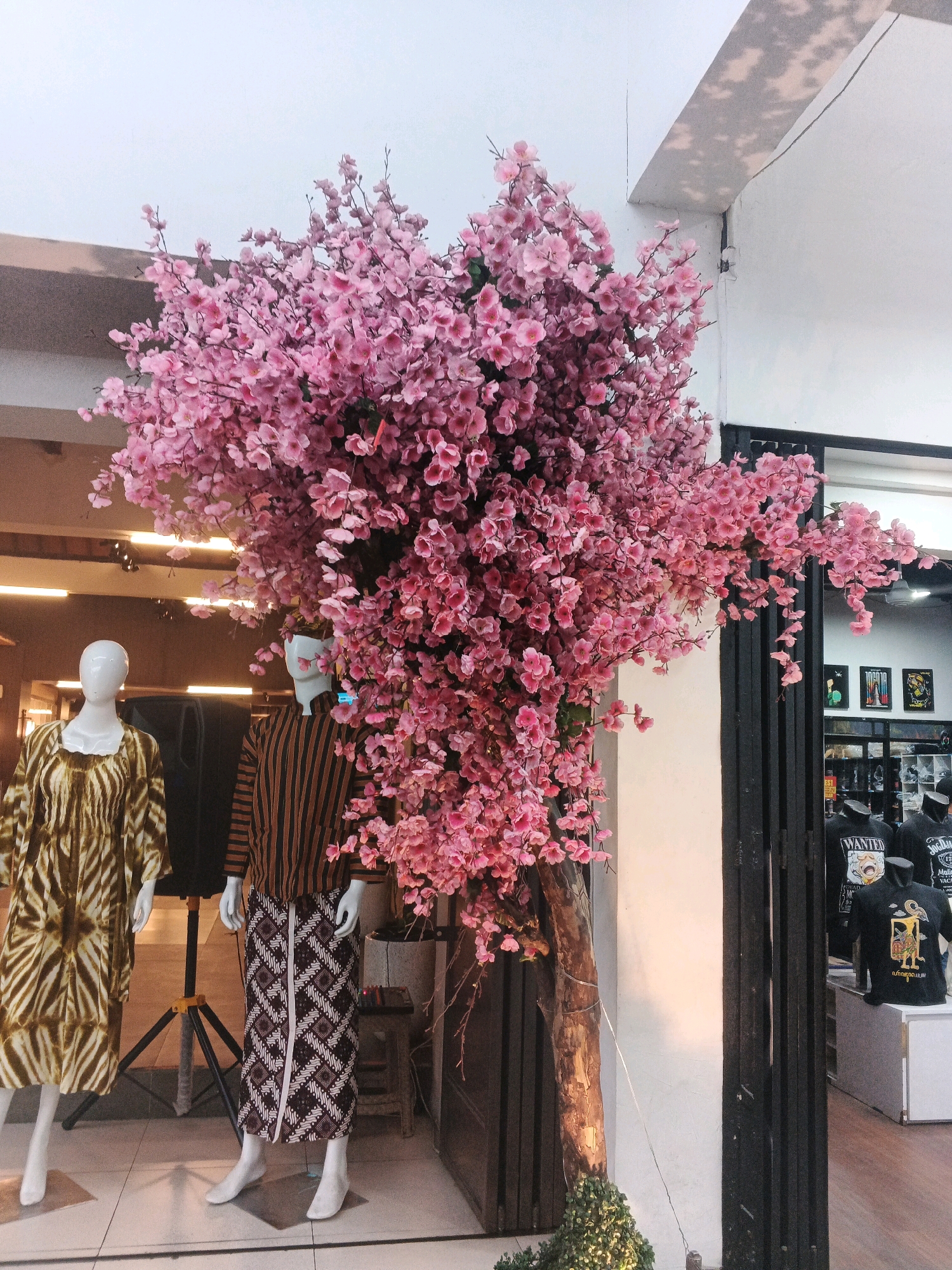 A tall, artificial pink cherry blossom tree dominates the center of the image, its branches cascading downwards. Two mannequins stand in front of the tree, positioned to the left of the frame. The mannequin on the left is wearing a light brown and white tie-dye dress. The mannequin on the right is dressed in a striped brown and black top and a patterned batik skirt. Behind the mannequins, a black speaker on a tripod is visible. To the right of the cherry blossom tree, a black metal gate with vertical bars is partially open, revealing a store interior. Inside the store, several black t-shirts with graphic designs are displayed on mannequins and shelves. The scene appears to be an outdoor or semi-outdoor display, possibly in front of a retail establishment in Yogyakarta, Indonesia, given the presence of batik clothing on the mannequin and the style of the store interior. The lighting suggests it is daytime, with a warm glow illuminating the tree trunk.