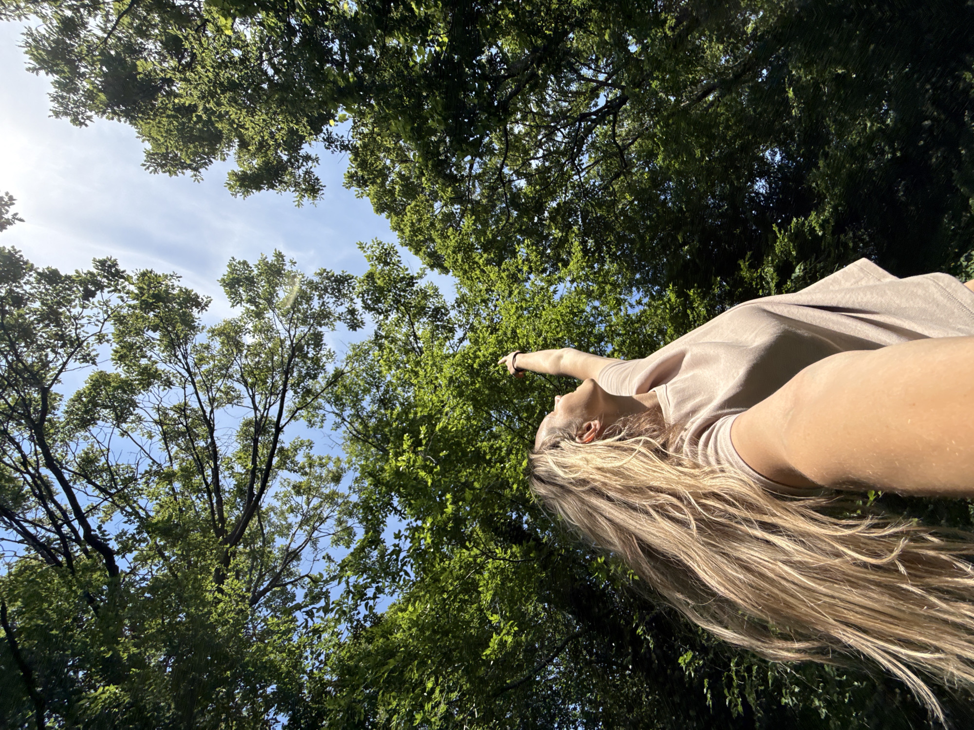 An outdoor, eye-level, medium shot shows a woman from the shoulders up with long blonde hair flowing behind her. The woman is looking up and to her right with her right arm extended upwards, her index finger pointing towards the sky. She is wearing a light brown t-shirt. The background consists of a canopy of lush green trees with a bright blue sky peeking through the leaves. The scene appears to be a forest or a park in the city of Nebug, Russia, under a sunny sky.