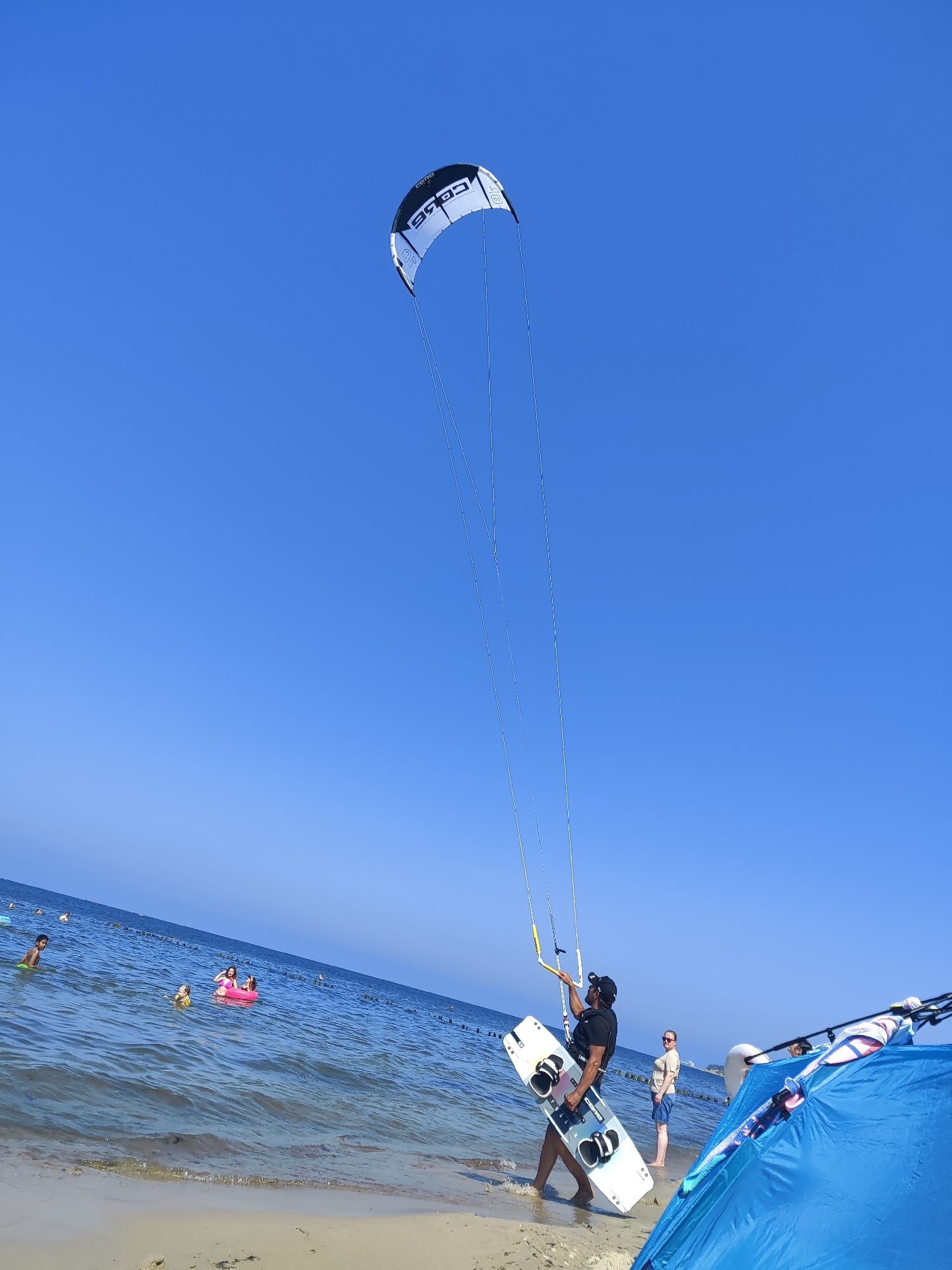 A group of people are enjoying a sunny day at the beach. The sky is a clear, bright blue, indicating good weather and likely daytime. In the foreground, a person is standing on the sandy shore, holding a white and black kiteboard. They are wearing a black t-shirt and shorts, and a cap. Another person, a woman, stands nearby in shorts and a t-shirt, looking towards the water. To the right, a blue beach tent is set up, with some equipment visible inside, possibly related to water sports.
In the background, the ocean stretches out to the horizon, with gentle waves lapping at the shore. Several people are in the water. Some are swimming, while others, including children, are playing with inflatable toys, like a pink ring. Wooden posts are visible in the water, likely forming a breakwater or marker. The scene appears to be a recreational beach setting. The kiteboard suggests activities like kitesurfing or kiteboarding may be taking place or about to take place. The overall mood seems relaxed and summery, with people enjoying a day by the sea. Text visible on the kite, partially obscured, reads "CORE". The location is Pionersky, Russia.