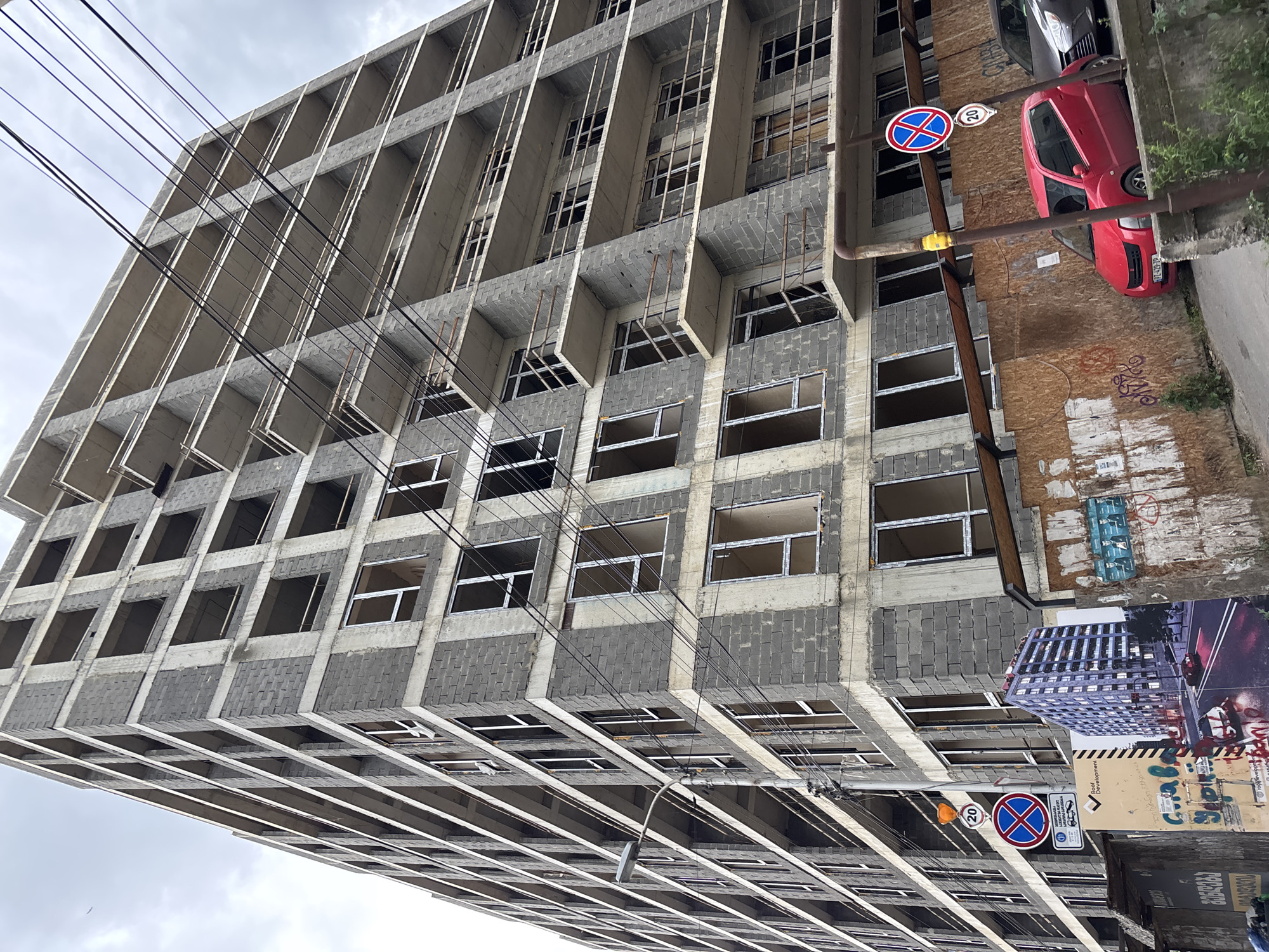 An upward view of an unfinished concrete building in Tbilisi, Georgia shows multiple window openings, many without glass, and concrete supports. Electrical wires run across the front of the building. A red car is parked next to the building near a sign with a red "X" through a blue circle and a speed limit sign of "20". A mural-like sign on the side of the building includes a picture of a finished version of the building.