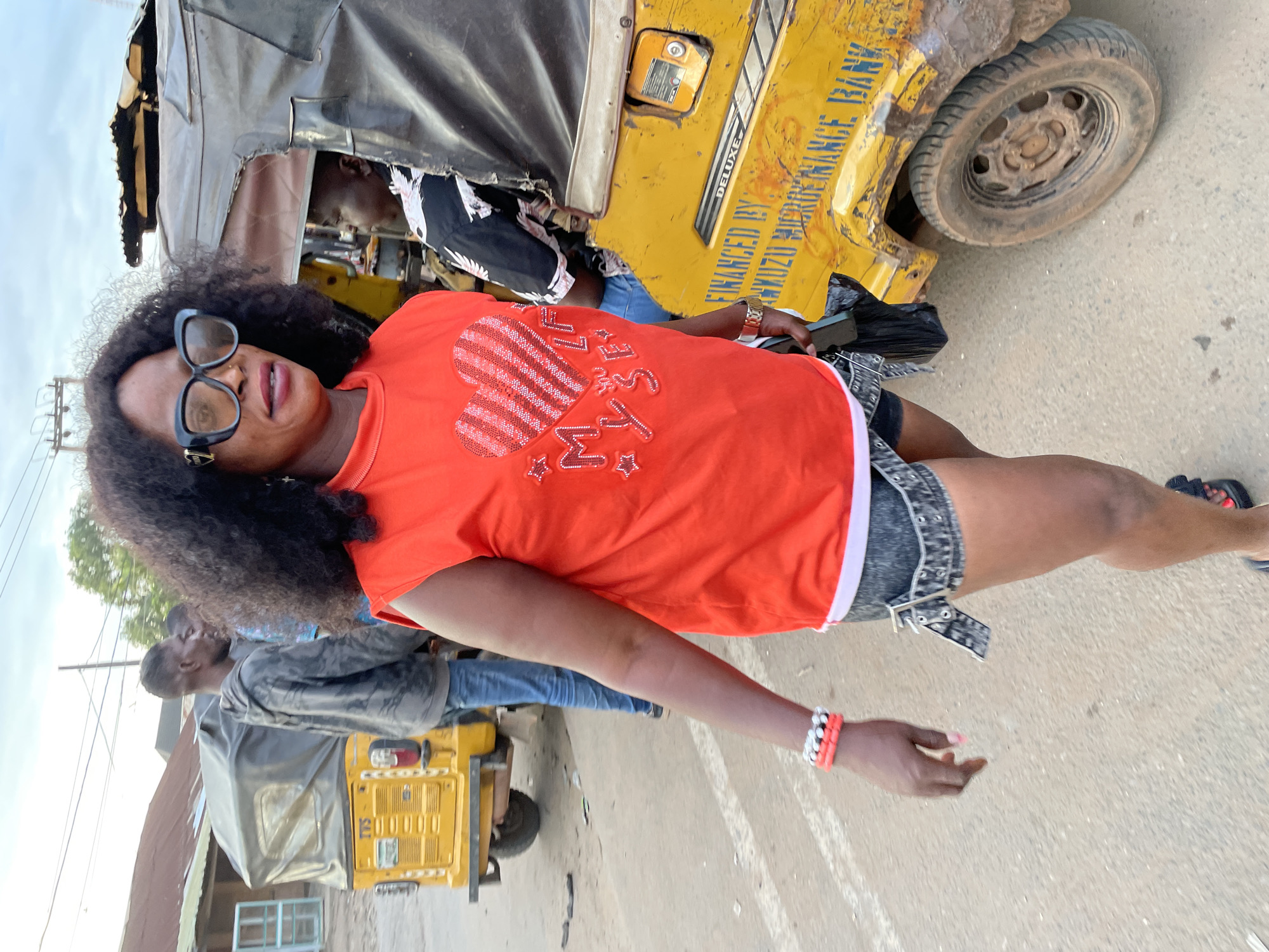 A Black woman in her 40s with dark, curly hair and dark sunglasses walks on a dirt road in Nkpor, Nigeria. She is wearing a sleeveless red t-shirt with a heart design and the words "MY LIFE" in glitter, denim shorts, and bracelets on her left wrist. She carries a black bag in her right hand. Behind her, a yellow auto-rickshaw with a black canopy is parked on the side of the road. A man wearing a gray hoodie and blue jeans is standing beside another auto-rickshaw in the background. The sky is cloudy and overcast. The road is dusty and appears to be a commercial area with buildings and shops visible in the distance.