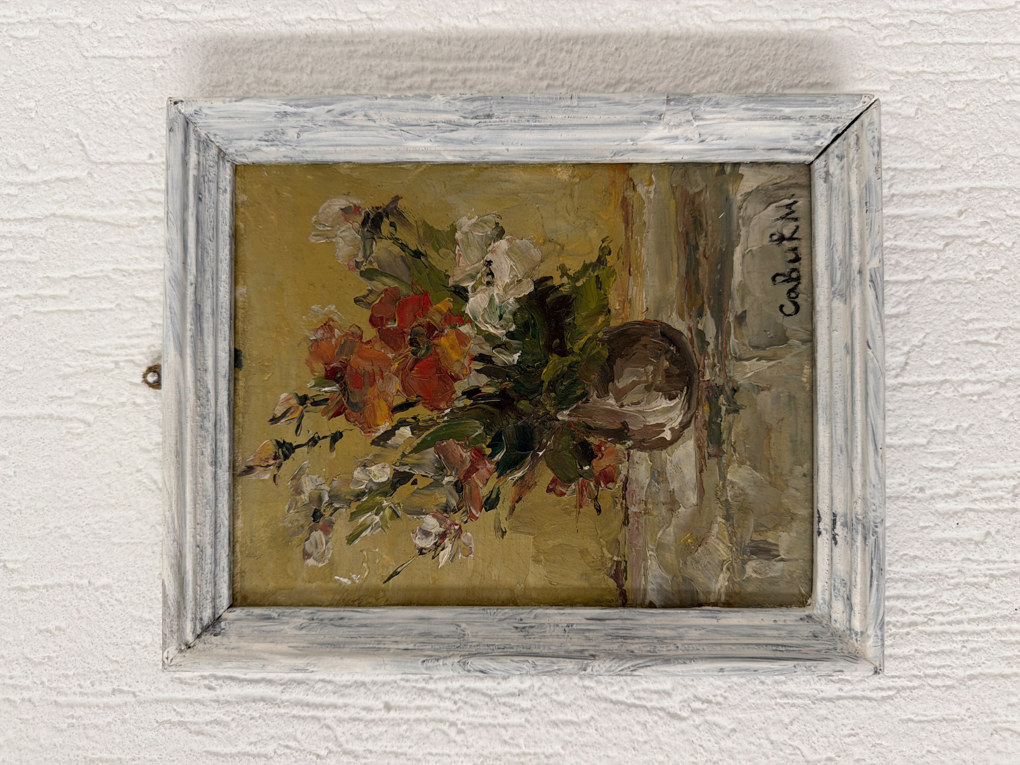 A framed painting hangs on a white, textured wall. The painting depicts a still life of flowers in a vase. The flowers are a mix of white, red, and orange hues. The background is a muted gold color. The vase is brown and appears to be slightly off-center to the left. The frame is ornate and painted white with gray undertones. The signature "Cabuk M." is visible on the right side of the painting. The location is not evident from the image.