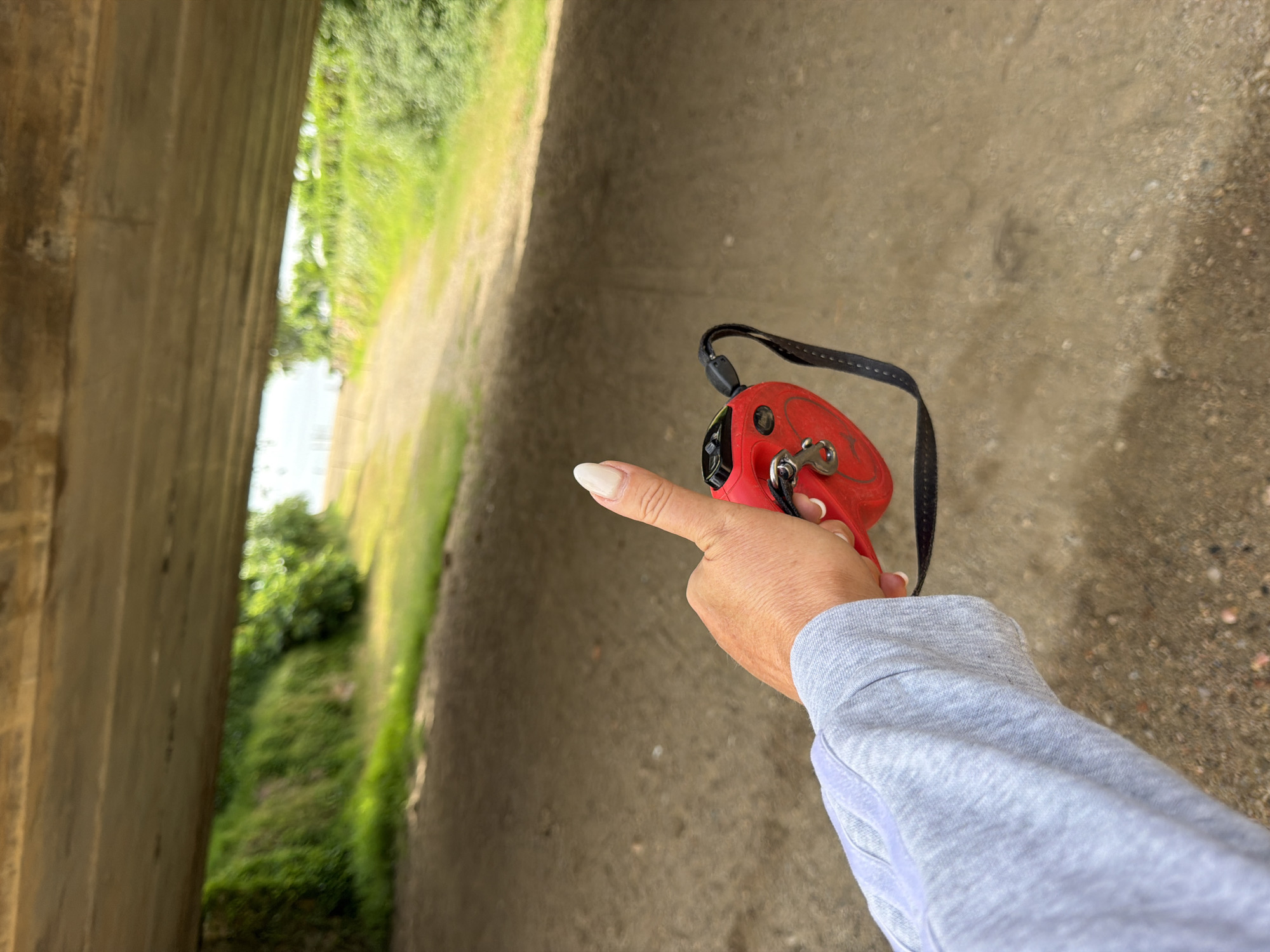 The image depicts a first-person perspective from beneath a concrete overhead structure, likely an underpass or bridge, situated in Mont-roig del Camp, Spain. In the lower-left quadrant, a light-skinned hand, presumably a right hand, is holding a red, oval-shaped retractable dog leash. The hand is covered by the sleeve of a light grey, long-sleeved shirt with a visible woven texture. The thumb is extended upwards, displaying a long, almond-shaped fingernail painted with a light, natural-toned polish. The red leash features a black fabric strap and a silver metal clip attached to its side. The ground beneath the concrete structure and extending forward is a light brown, unpaved surface consisting of dirt and small pebbles. Beyond the structure, a dirt path or road leads into the distance, flanked on both sides by dense green vegetation, including bushes and trees. The background beyond the foliage appears hazy and brightly lit, possibly indicating open sky or distant water.