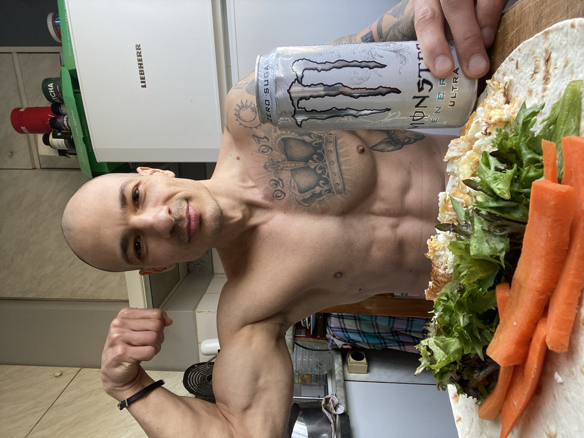 The scene captures an adult male, shirtless, in a domestic kitchen setting located in Pleven, Bulgaria. The individual, possessing a shaved head and light skin, stands with his left bicep flexed towards the viewer. His right hand holds a silver "MONSTER ENERGY ULTRA ZERO SUGAR" can. The man exhibits a muscular physique, with visible abdominal muscles, chest, and arms. Prominent tattoos include a large crown on his chest with the numbers "02" and "07" positioned above it, a sun and moon design on his left shoulder, and additional imagery on his left arm and lower left torso. He wears a black wristband on his left arm and has a slight smile.
In the immediate foreground, a meal rests on what appears to be a wooden surface, likely a cutting board. The meal consists of a flatbread or tortilla topped with mixed green lettuce and a scrambled egg-like preparation. Alongside this, several peeled and sliced raw carrots are arranged.
The background displays elements of a kitchen. A white "LIEBHERR" brand refrigerator is situated behind the man's right side. Above the refrigerator, a green box containing "DRINK BETTER FEEL BETTER" and "MATCHA" is visible, along with a red cylindrical container and other supplement jars, one labeled "L-CARNITIN." To the man's left, a light-colored countertop holds a partially visible black fan, a white small appliance, a clear plastic bottle, and a plaid fabric. Light-colored wall tiles or panels and upper cabinets with a light wood finish complete the background.