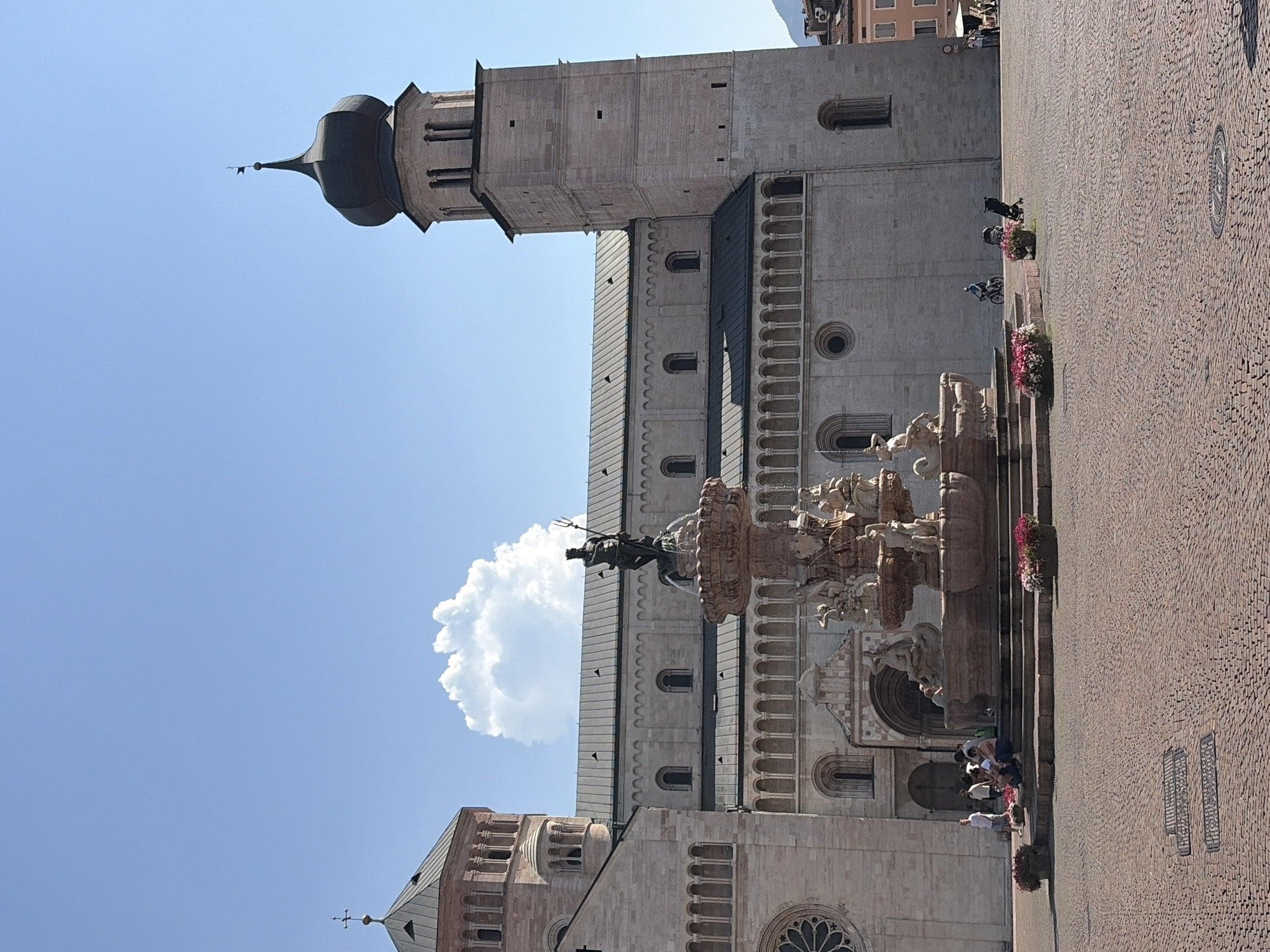 The image shows Piazza Duomo in Trento, Italy. A large, ornate fountain with a statue of Neptune stands in the foreground. Behind the fountain is the Cathedral of San Vigilio, a Romanesque cathedral with two bell towers. The sky is clear and blue with a single white cloud. Several people are gathered in the piazza, some sitting on the steps of the fountain, others walking or standing around. A stroller and a bicycle are also visible in the scene. The ground is paved with cobblestones.