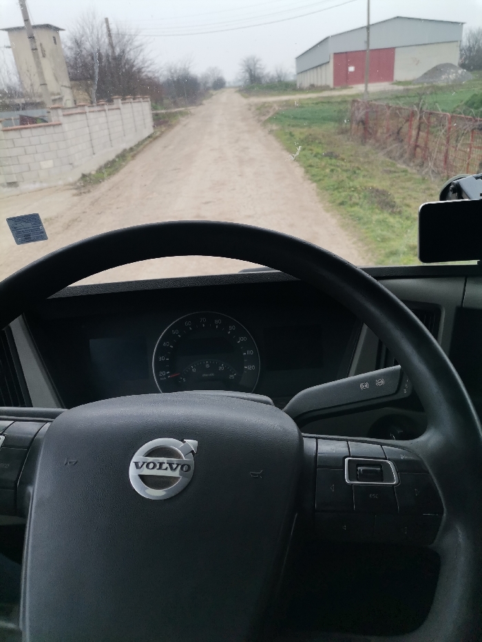 The image presents a first-person view from the driver's seat of a Volvo vehicle, situated on an unpaved dirt road in Kardam, Bulgaria. The black Volvo steering wheel, bearing the silver Volvo logo, dominates the lower foreground, with dashboard instruments including a speedometer and tachometer visible behind it. A smartphone is mounted on the right side of the dashboard. A rectangular sticker is affixed to the lower-left of the windshield, which shows minor smudges.
The dirt road, marked by tire tracks, extends centrally into the distance under an overcast sky. To the left, a low concrete block wall parallels the road, behind which stands a tall, light-colored concrete utility structure and sparse, bare trees. To the right, green vegetation and a rusty metal fence border the road. In the mid-ground right, a large, light-colored industrial-style building with a distinct red section is visible, fronted by a pile of aggregate and a utility pole.