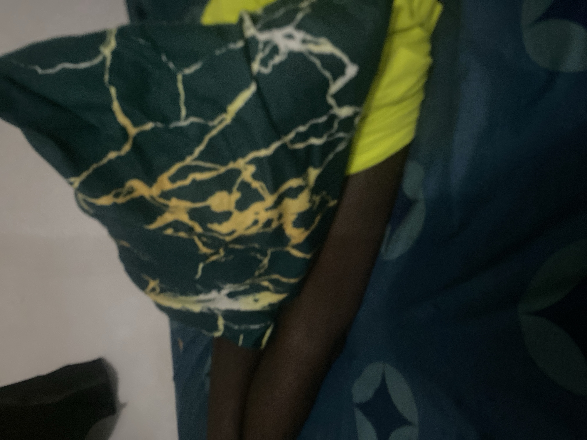 The image shows a close-up view of a person's legs, covered partially by a dark green pillow with a yellow and white marbled pattern. The person is lying on a dark blue bedsheet with a subtle geometric pattern. A bright yellow garment is visible underneath the pillow and partly covering the legs. The background is a plain, light-colored wall, with a dark object hanging in the upper left corner. The scene appears to be indoors, likely in a bedroom in Howayi, Nigeria. The lighting is dim, suggesting it might be nighttime or the room is not well-lit.