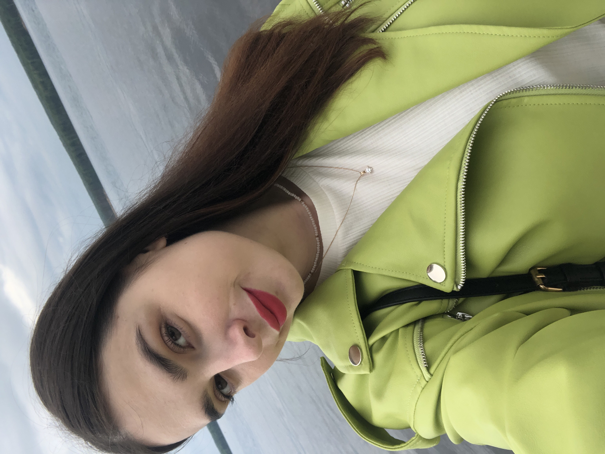 A young woman with long, dark brown hair, light brown eyes, and red lipstick poses for a selfie outdoors. She is wearing a bright lime green leather jacket over a white ribbed top. A delicate necklace with small pearls and a thin gold chain with a small pendant are visible. A black strap with a brass buckle is draped across her chest. The background consists of a body of water, likely the Kama River, with a distant, tree-lined shore under a cloudy sky. The photographer is positioned close to the subject, capturing her face and upper body. The scene appears to be in Chaikovsky, Russia.