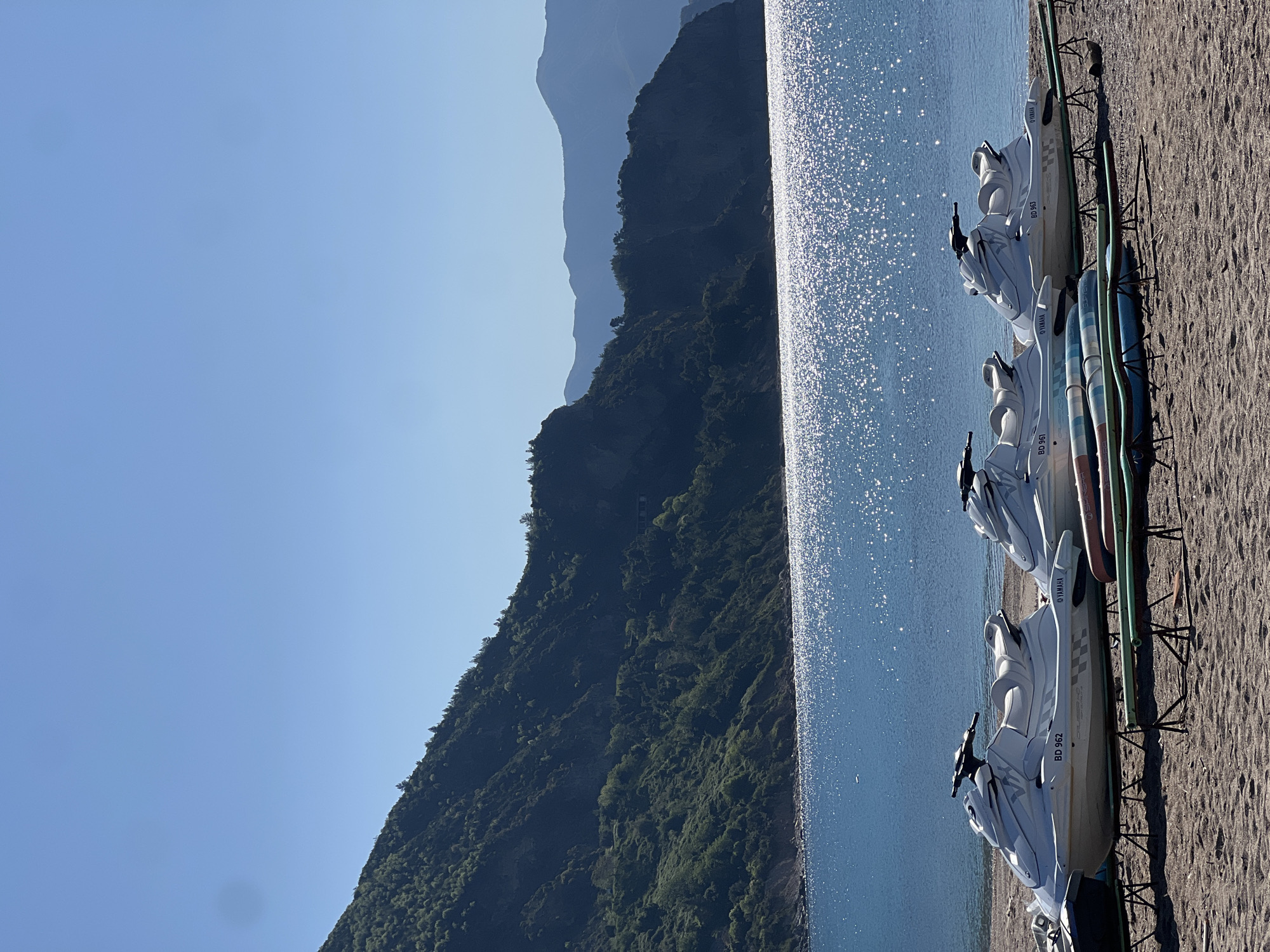 A vibrant landscape image, captured on a sunny day in Prijevor, Montenegro, depicts a beach scene next to a body of water with dramatic mountain backdrops. In the foreground, on a sandy shore, several white jet skis are neatly parked on green-colored stands, facing the water. The jet skis are branded "YAMAHA" and display the registration number "BD 962" on their sides. Alongside them, multiple paddleboards or surfboards are also visible, resting on similar racks.
To the right, a large expanse of water glistens brightly, reflecting the sunlight in numerous sparkling points. In the background, a series of towering mountains, densely covered in green foliage, rise steeply from the water's edge. Further distant mountain ranges appear progressively lighter and bluer, creating a sense of depth and scale against a clear, bright blue sky.
The clear sky and intense water sparkle indicate bright, sunny weather during daytime. The presence of jet skis and paddleboards suggests a recreational area offering water sports, evoking feelings of leisure and outdoor activity in a scenic natural setting. There are no people visible in the image.