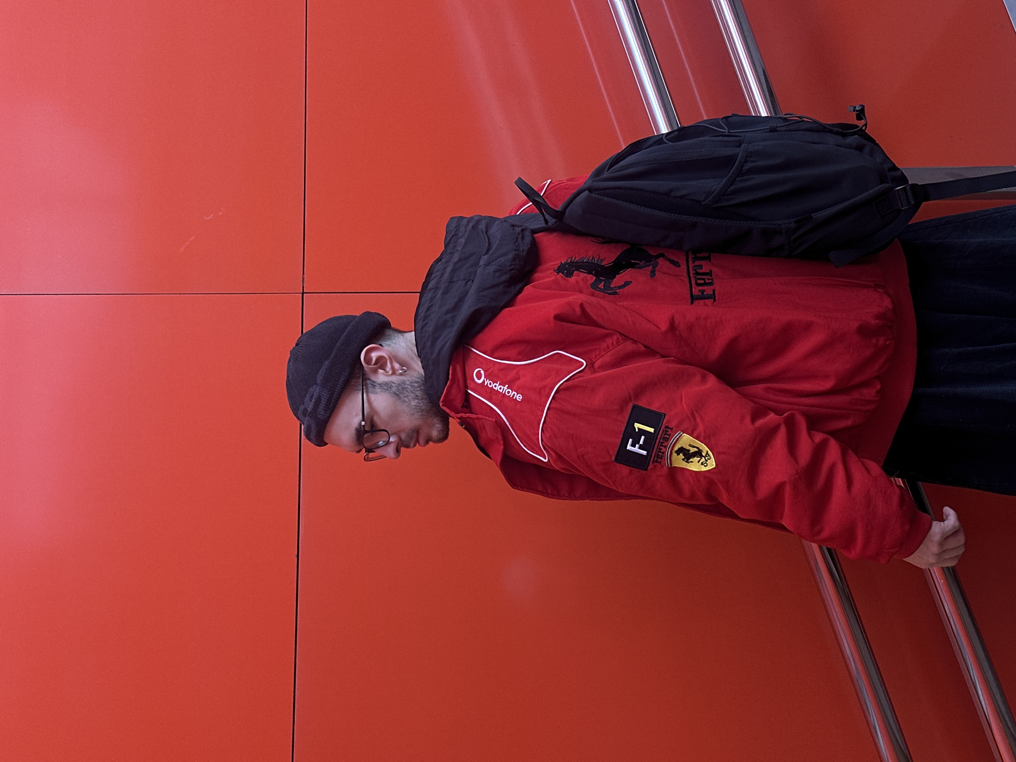 The image features a man in profile, oriented diagonally, against a striking orange-red wall. He is wearing a black beanie, eyeglasses, and a vibrant red racing jacket. A black backpack is worn over his shoulders, and his head is tilted downwards. A small silver hoop earring is visible on his left ear.
The red jacket prominently displays a large black Ferrari prancing horse logo on the back. On the right sleeve, there are multiple patches, including a black and white "F-1" emblem and a yellow Ferrari shield logo featuring the black prancing horse and "Ferrari" text. The "Vodafone" logo in white is visible on the front chest area of the jacket.
Behind the man is a clean, segmented orange-red wall. Two parallel, polished silver metal railings run diagonally in the foreground, suggesting he is on an escalator or staircase. His right hand is lightly gripping one of these railings. The lighting is even, indicating an indoor environment. The setting for this scene is Moscow, Russia.